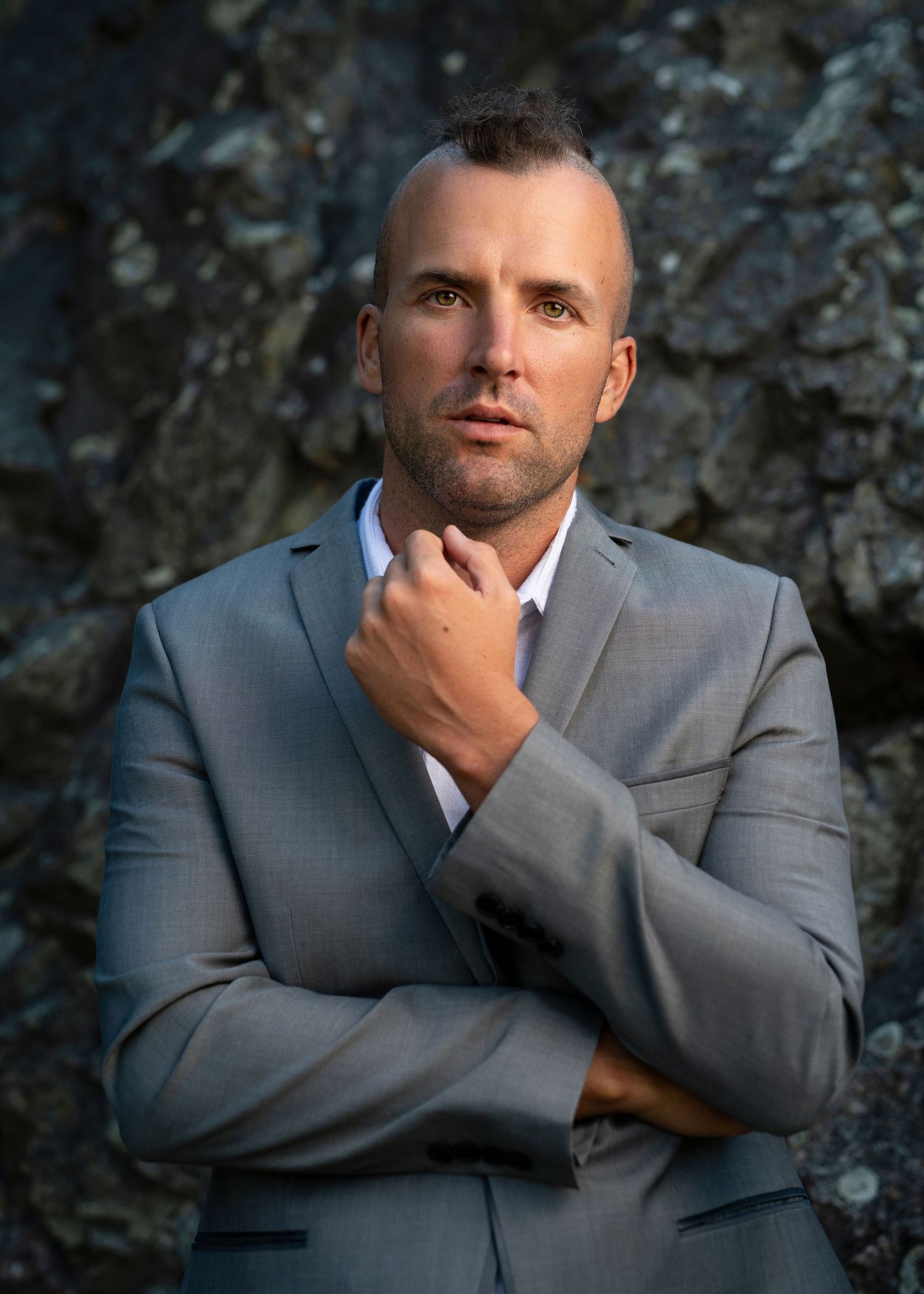Young man in grey suit posing in front of rock wall with hand raised near chin, looking at camera - Okanagan portrait by Tania Veitch.