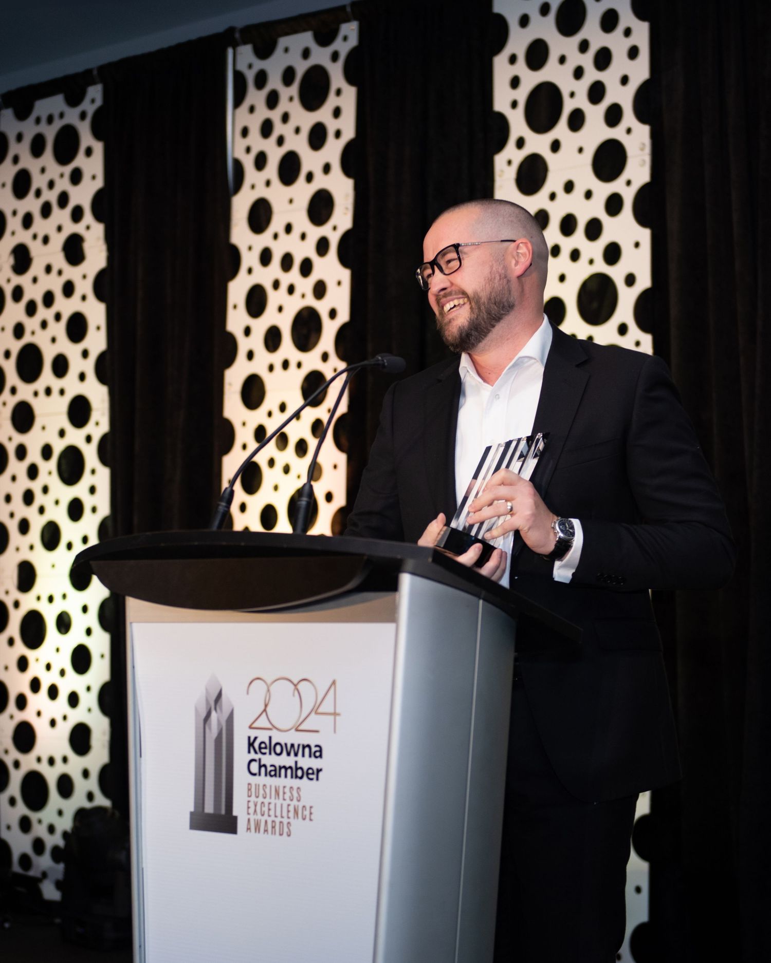 Young man with beard and glasses holding award on stage at Kelowna Chamber Business Excellence Awards - photo by Tania Veitch.