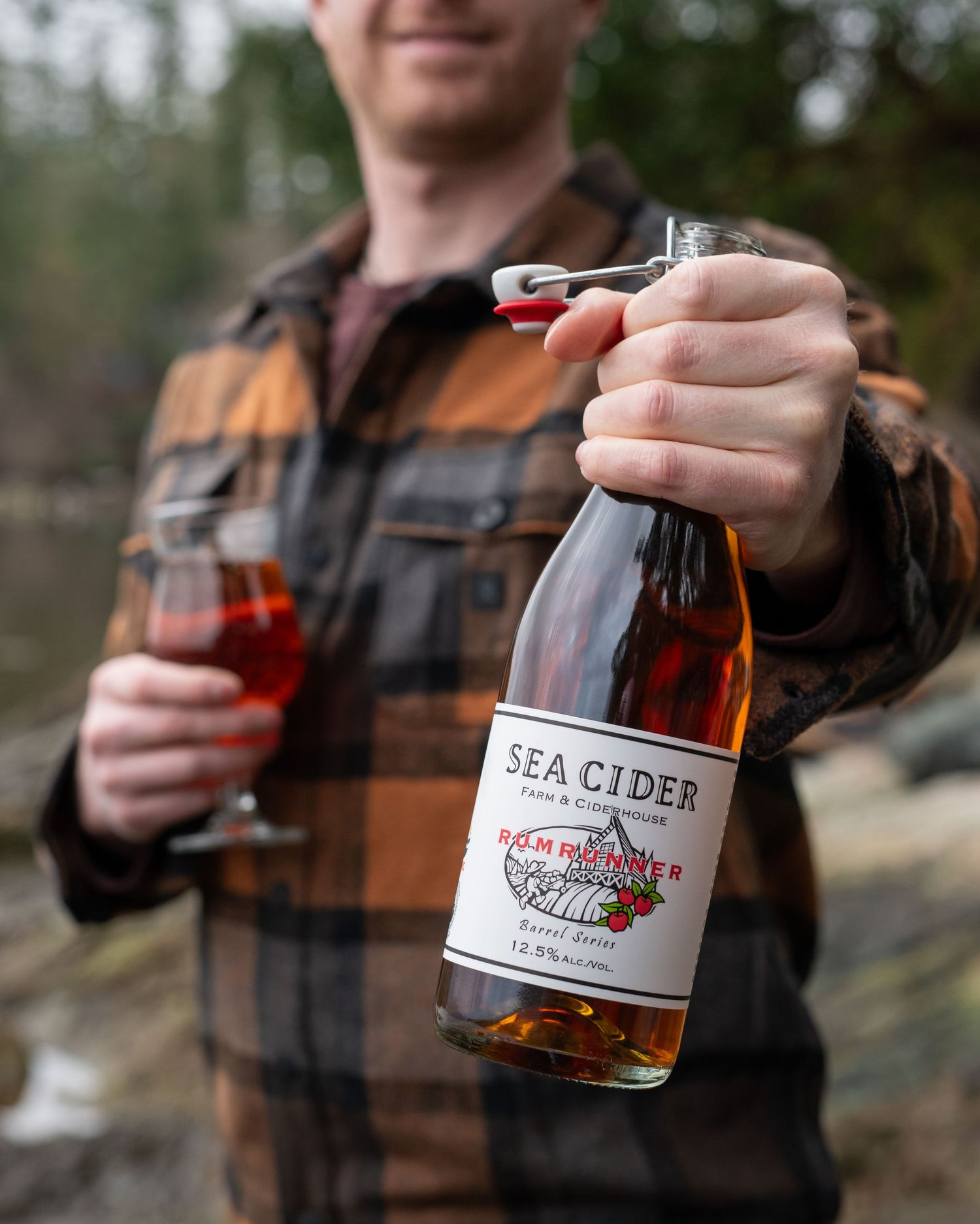 Man in plaid shirt holding Sea Cider Rumrunner bottle toward camera, a full glass in his other hand - Okanagan brand photo by Tania Veitch.
