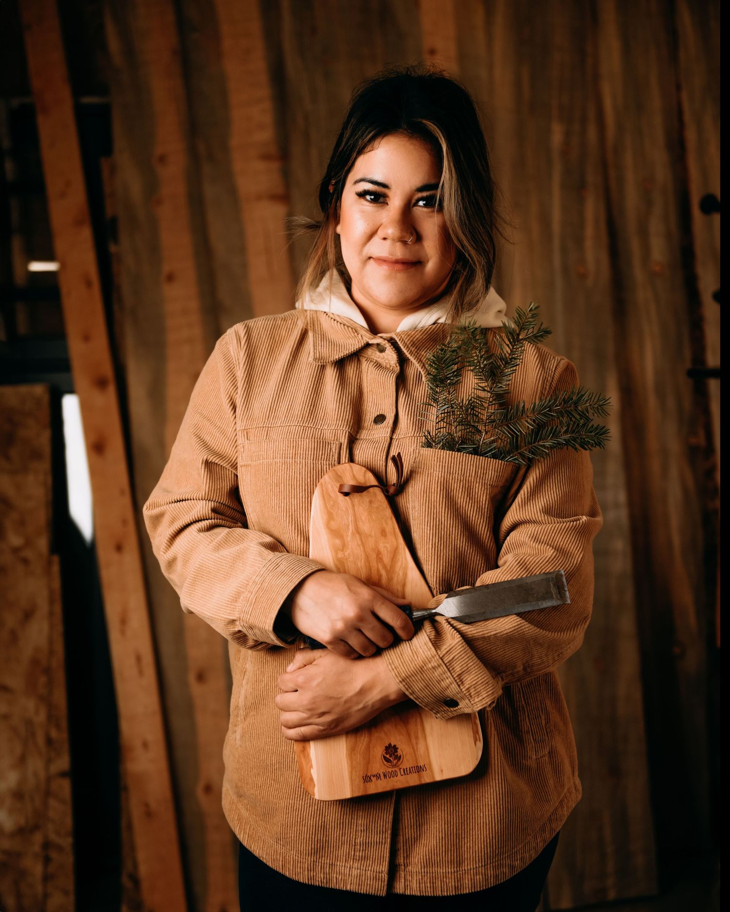 Young First Nations woman in tan jacket holding charcuterie board and chisel before stacked boards - Okanagan portrait by Tania Veitch.