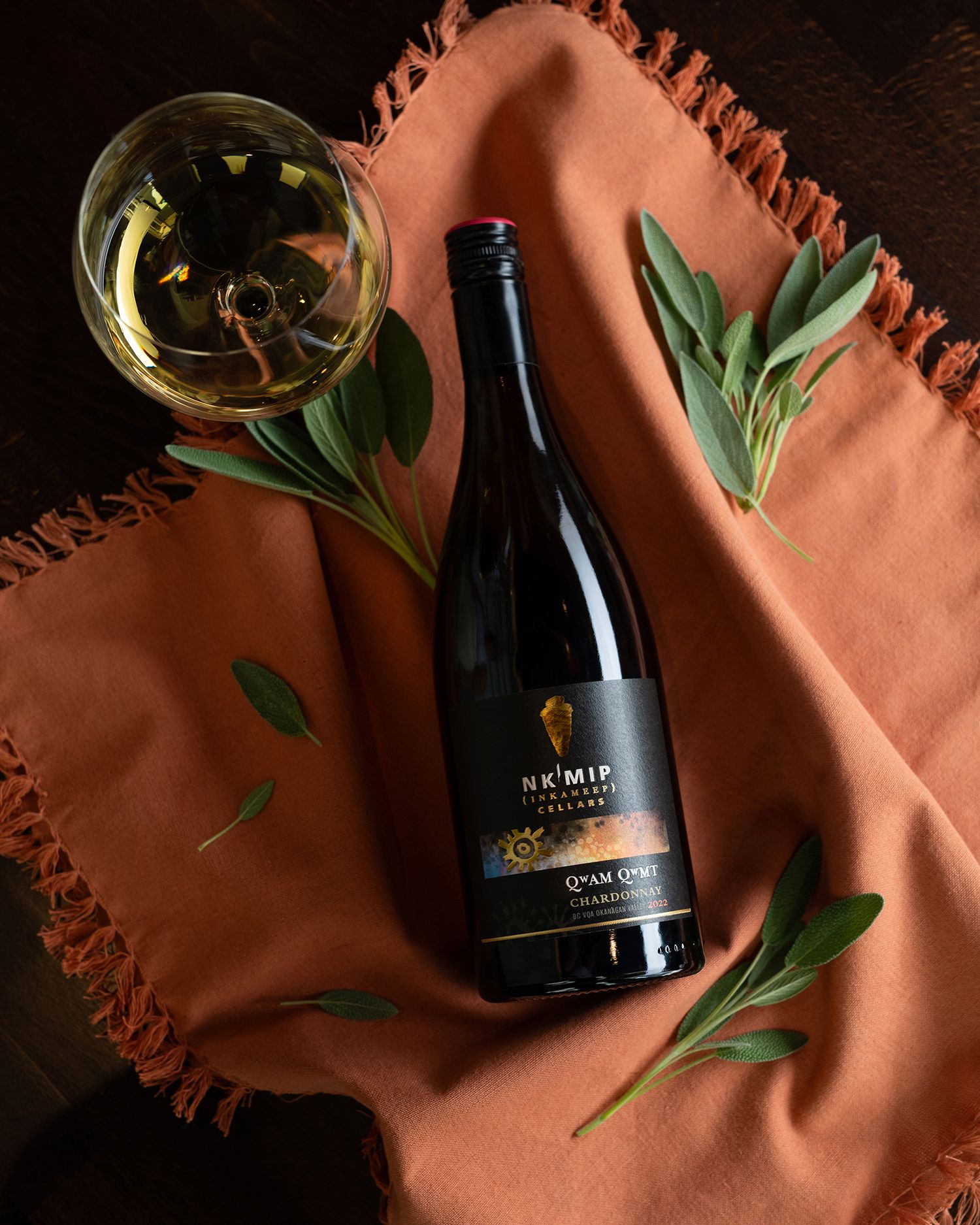 Styled flat lay of Nk’Mip Cellars Chardonnay bottle with wine glass and sage on textured linen background.