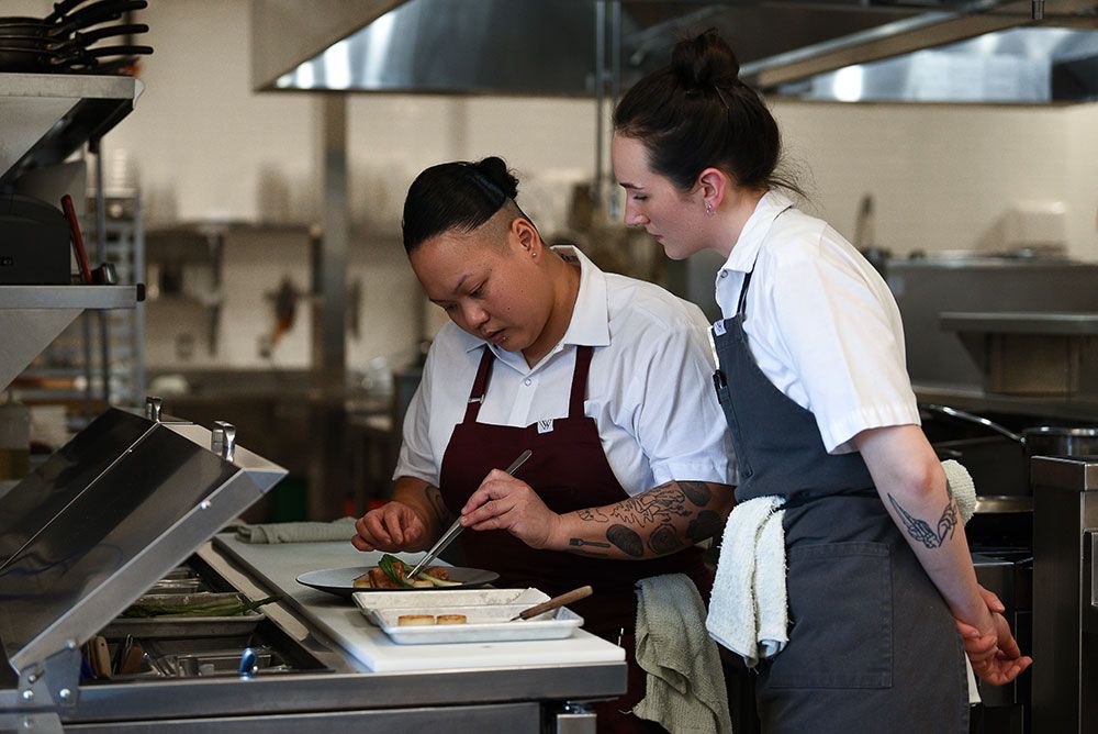 Restaurant brand photography of chef plating a gourmet dish while assistant chef observes in professional kitchen.