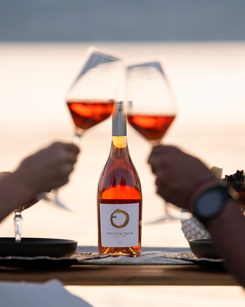 Phantom Creek rose wine bottle on table by lake with two people clinking glasses, Okanagan wine photo by Tania Veitch.