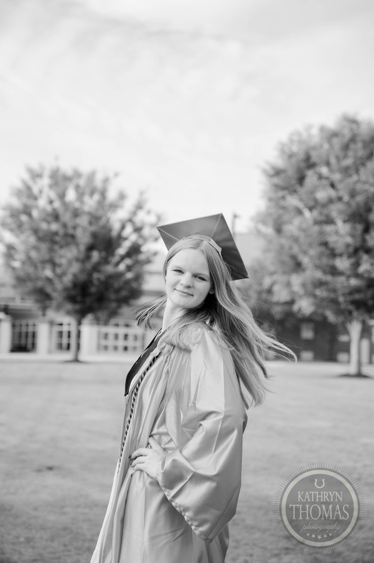 Graduate in cap and gown poses outdoors on campus grounds during graduation photoshoot with trees in background.