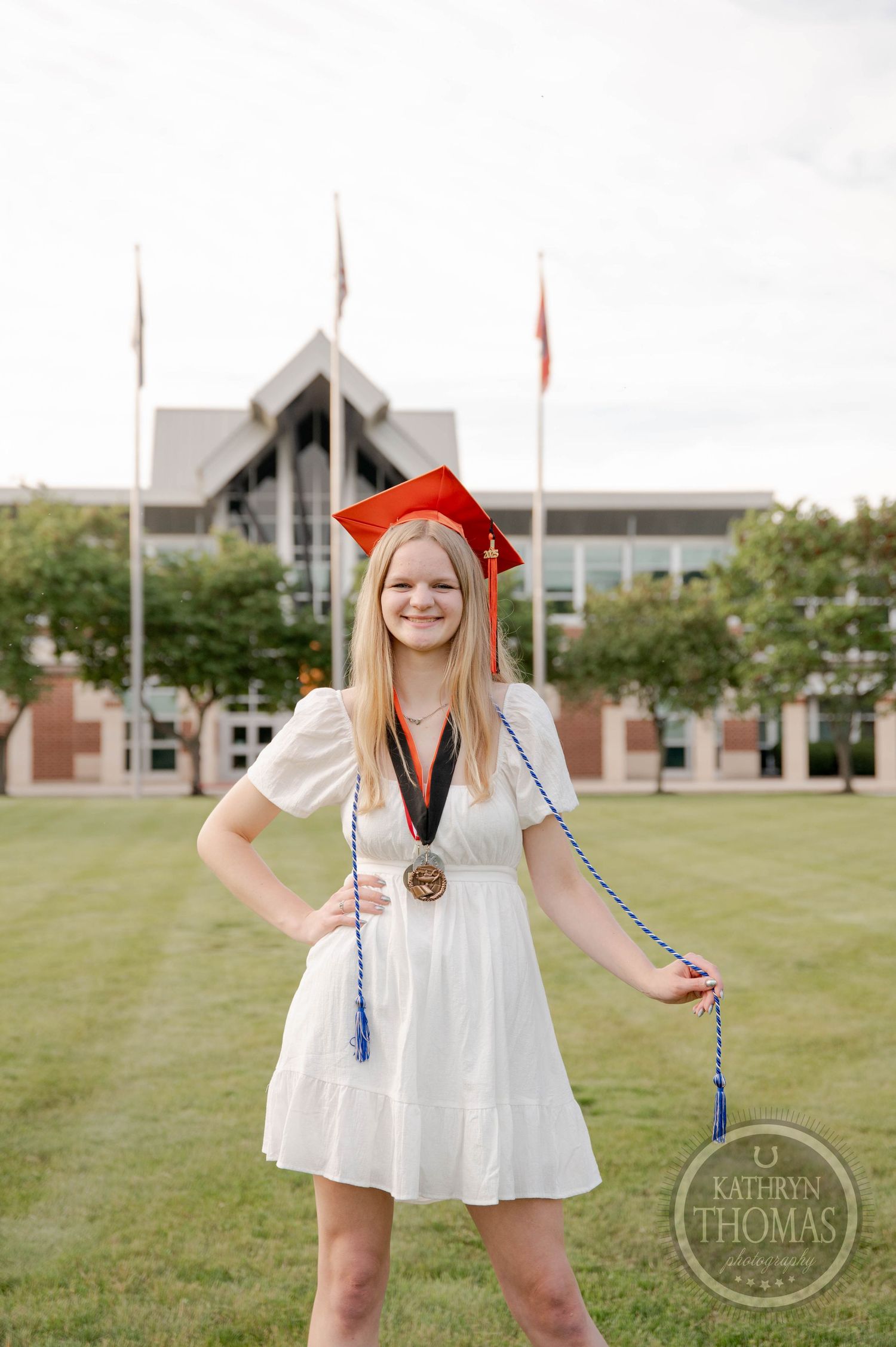 College graduate stands outdoors in white dress and red cap with campus buildings visible in the background.