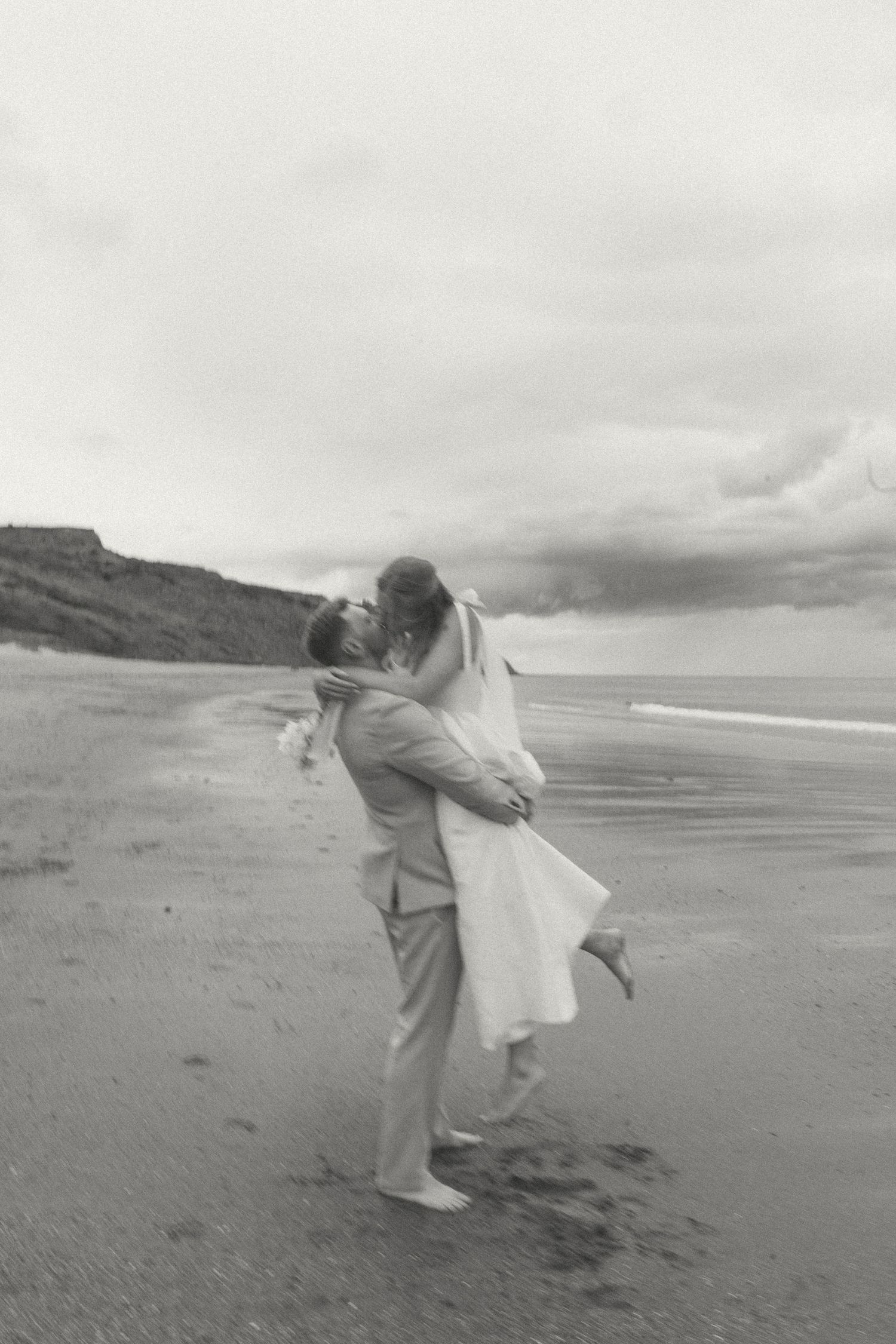A black and white analogue image showing a couple embacing on a windswept beach with cloudy skies.