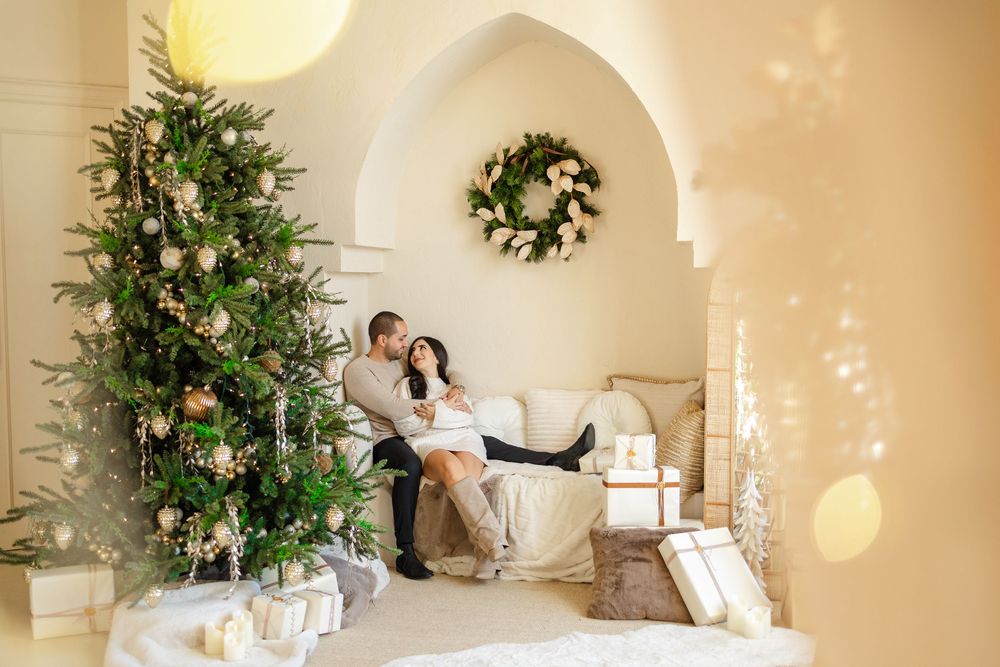 A couple sits in a white archway surrounded by a decorated Christmas tree, wreath and wrapped presents.
