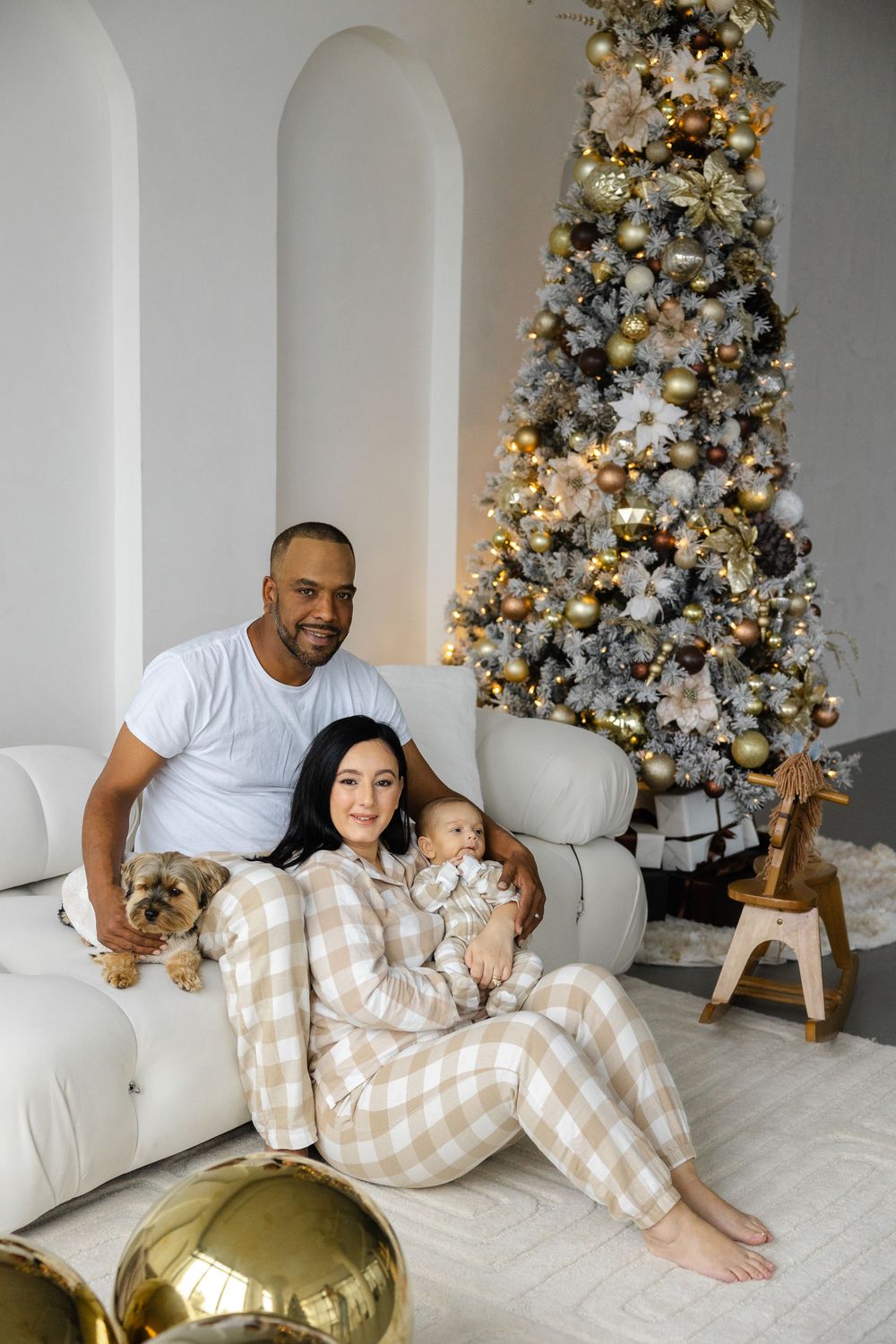A family in matching plaid pajamas sits together on a white couch beside an elegantly decorated Christmas tree.
