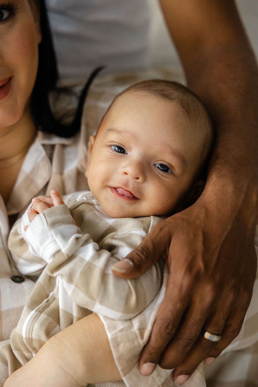 A baby wearing a white outfit lies on soft bedding while being comforted by gentle hands.