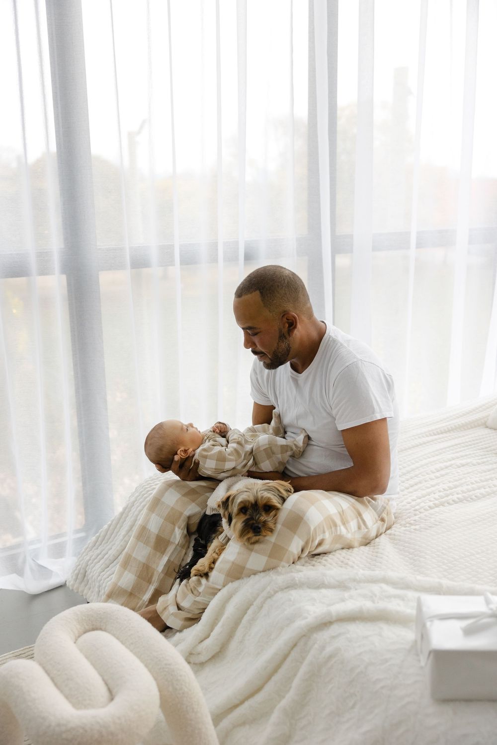 A tender moment as a man in white embraces someone covered in soft blankets by sunlit windows.