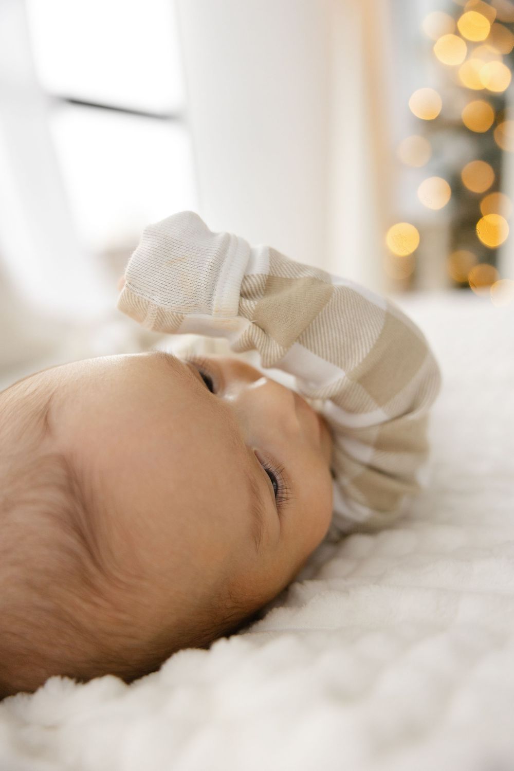 A sleeping baby lies peacefully on white bedding with soft bokeh Christmas lights glowing in the background.