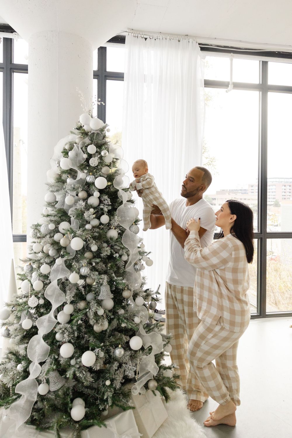 Three decorative white Christmas trees are arranged in an interior space while decorators add elegant white ornaments and decor.
