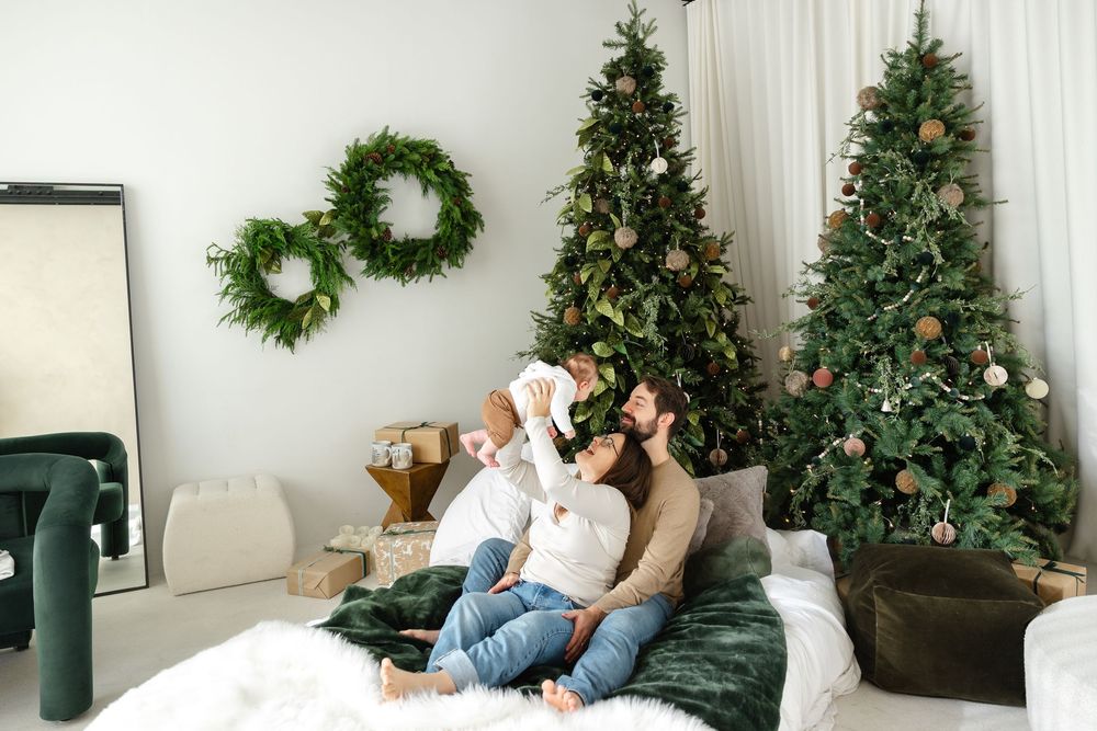 A cozy holiday scene with two Christmas trees, wreaths on the wall, and people relaxing on a white couch together.