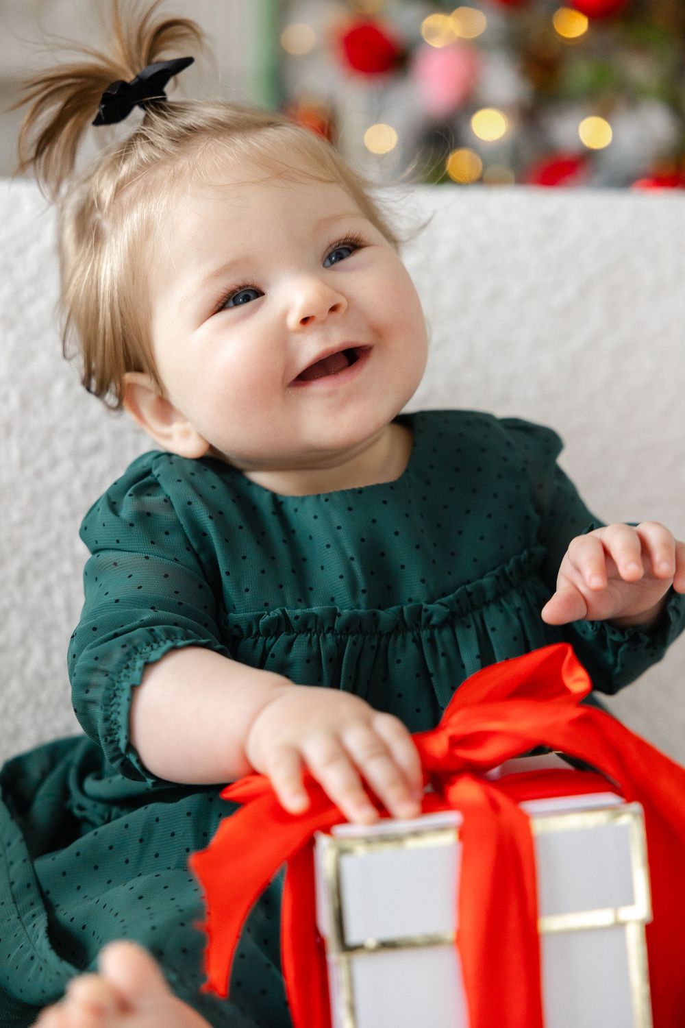 Baby in emerald green dress smiles joyfully while holding Christmas present with red bow against blurred holiday lights.
