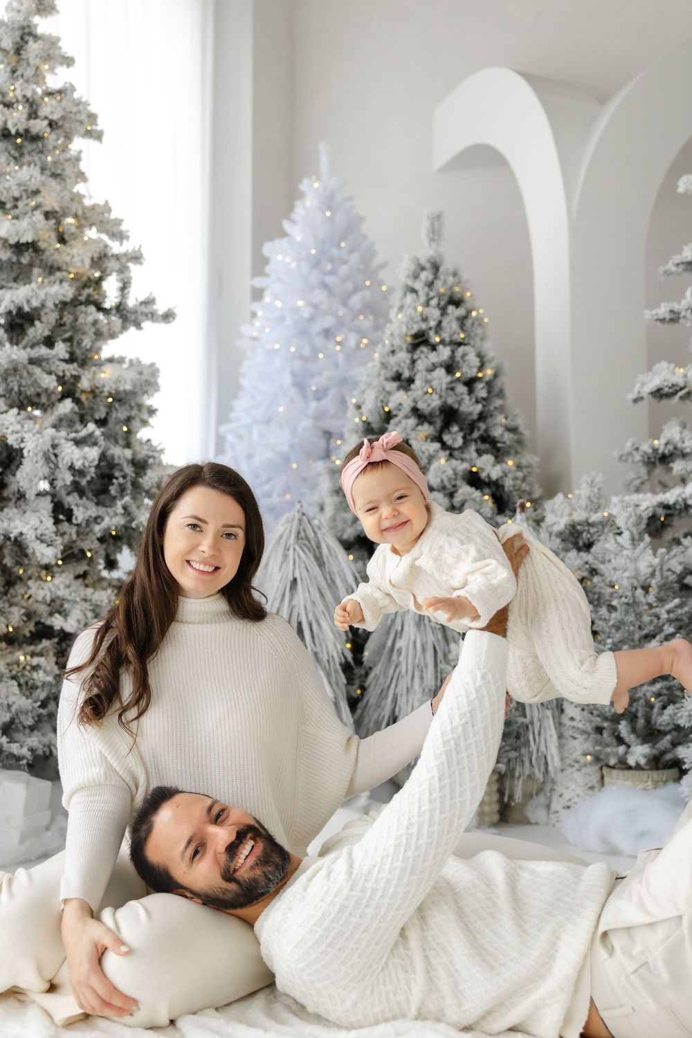 Family in cozy white sweaters pose playfully in front of decorated white Christmas trees in a bright holiday photoshoot setting.