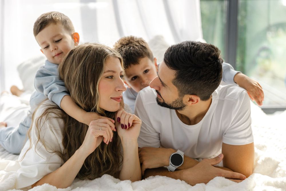 Family relaxing together on white bedding in bright morning light.