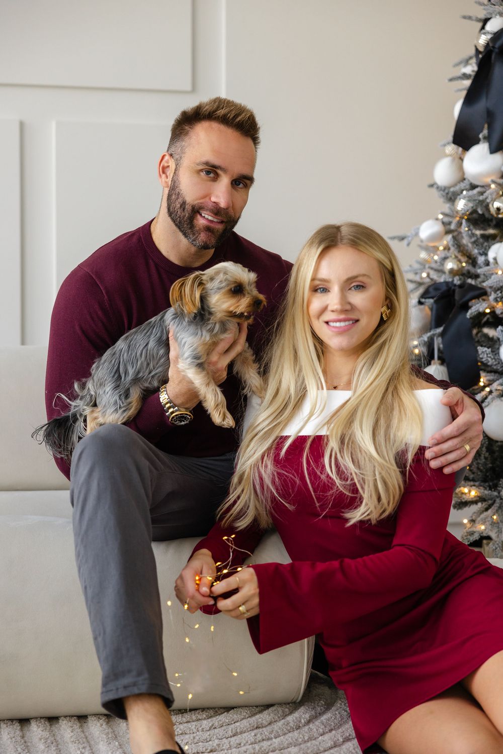 Festive holiday portrait of a couple in burgundy outfits posing with their small terrier dog next to a Christmas tree.