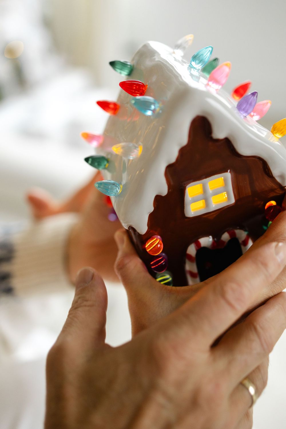 Four sequential photos show hands holding a handmade gingerbread house decorated with colorful candies and white frosting.