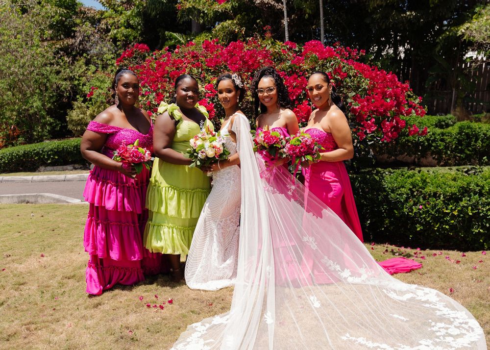 Bridal party in pink and white dresses pose together outdoors with flowering bushes in background.