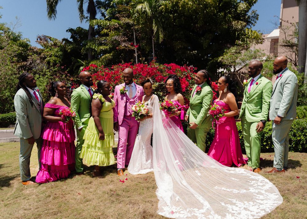 Wedding party members in coordinating bright pink and lime green attire pose outdoors in a garden setting.