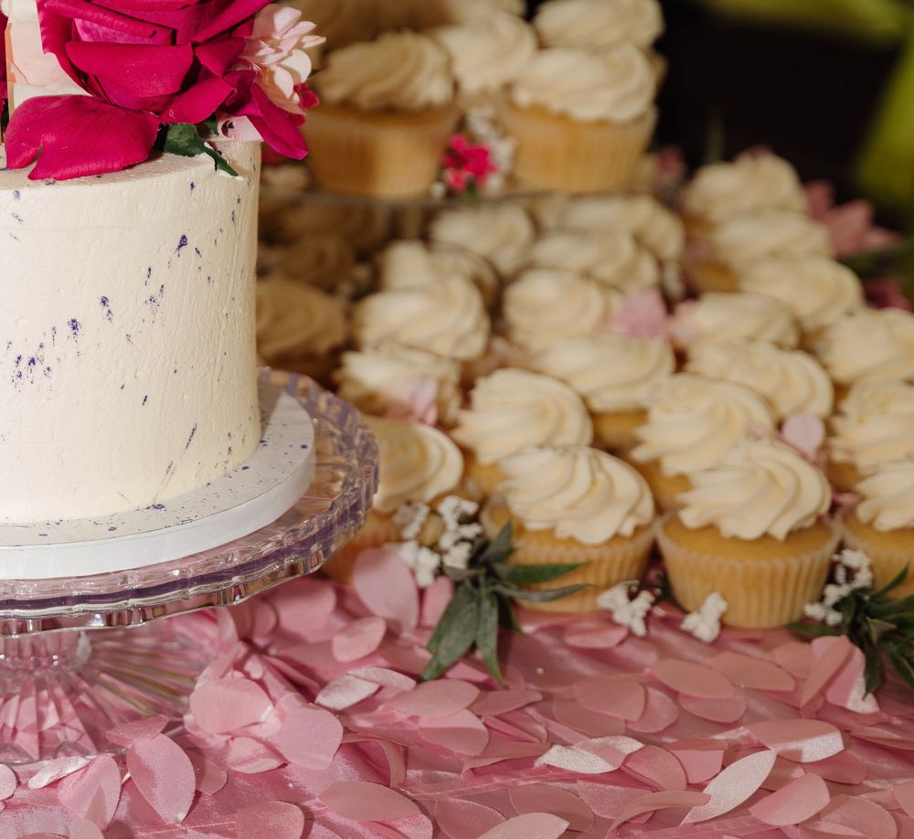 A festive display of white cupcakes arranged around a pink and white tiered wedding cake with floral decorations.