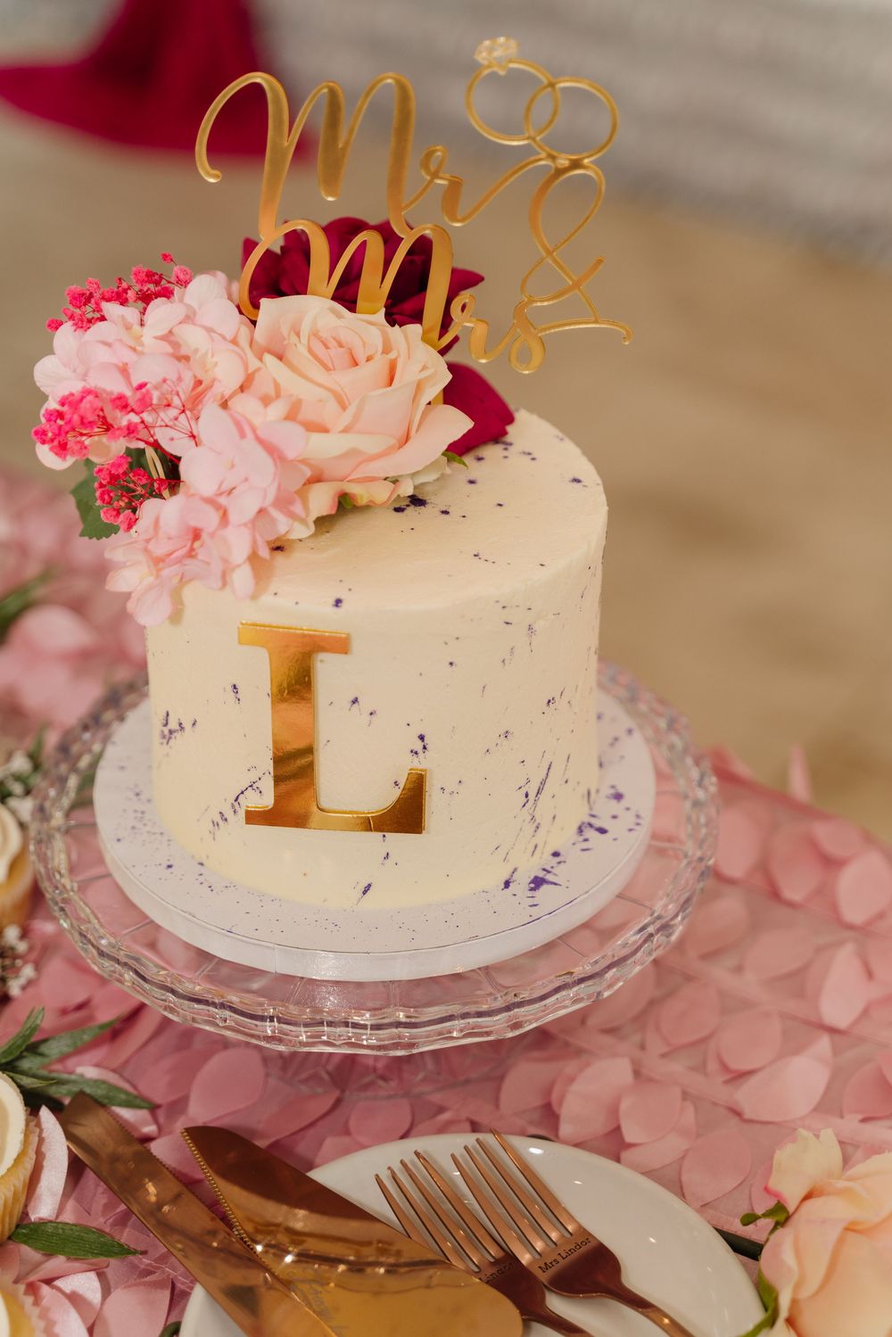 White wedding cake decorated with pink roses and gold accents on a glass stand.
