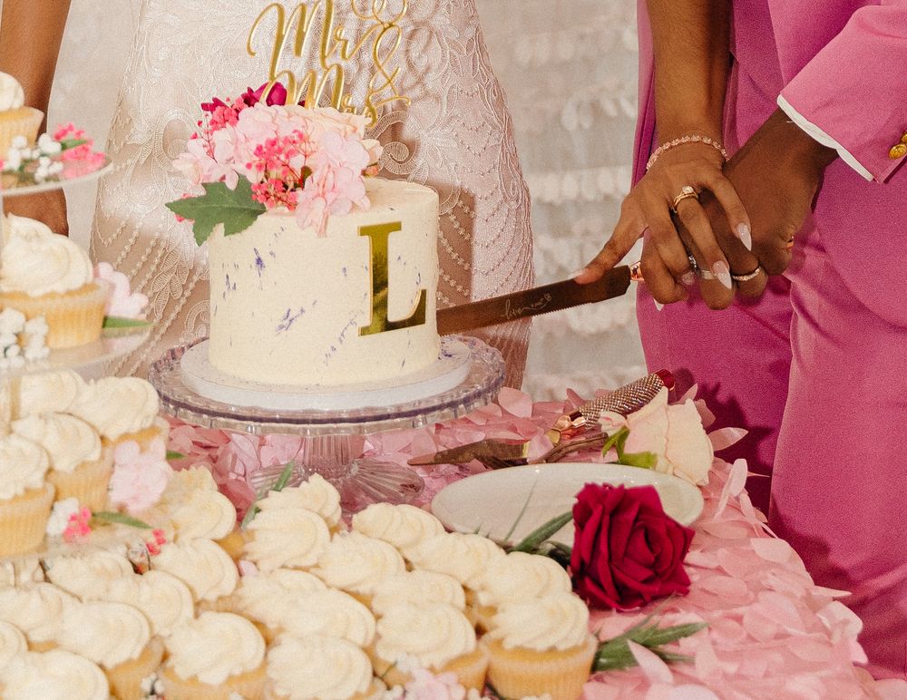 A beautifully decorated white wedding cake with pink floral details and gold monogram being cut beside pink and white desserts.