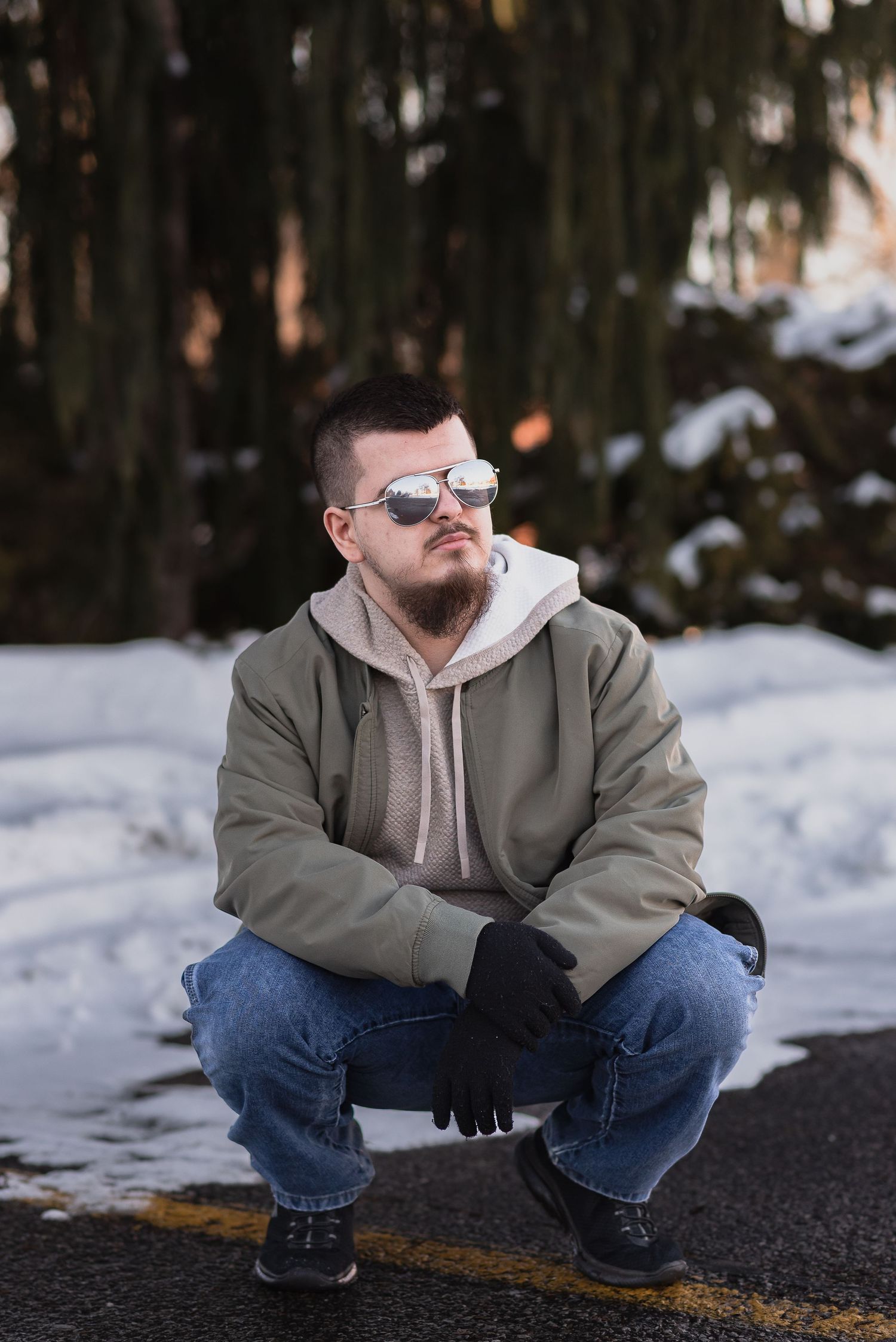 Man in olive jacket, hoodie, jeans, and mirrored sunglasses crouching on a road in a snowy winter setting.