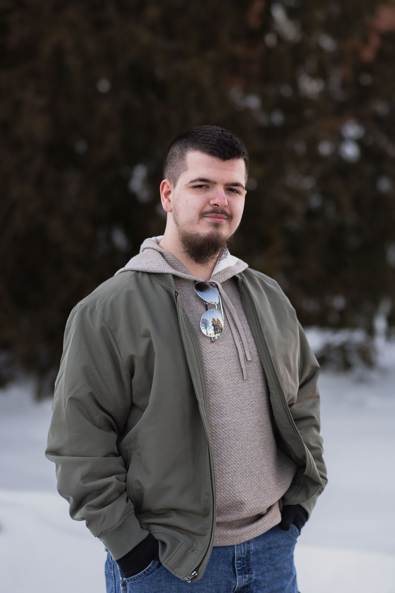 Young man with beard wearing olive jacket and hoodie, standing outdoors in snowy winter landscape.