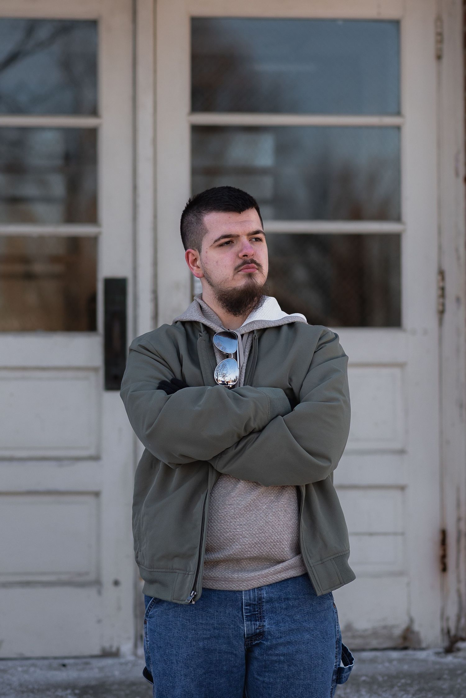 Young man with beard in olive jacket, arms crossed, standing confidently in front of vintage white double doors.