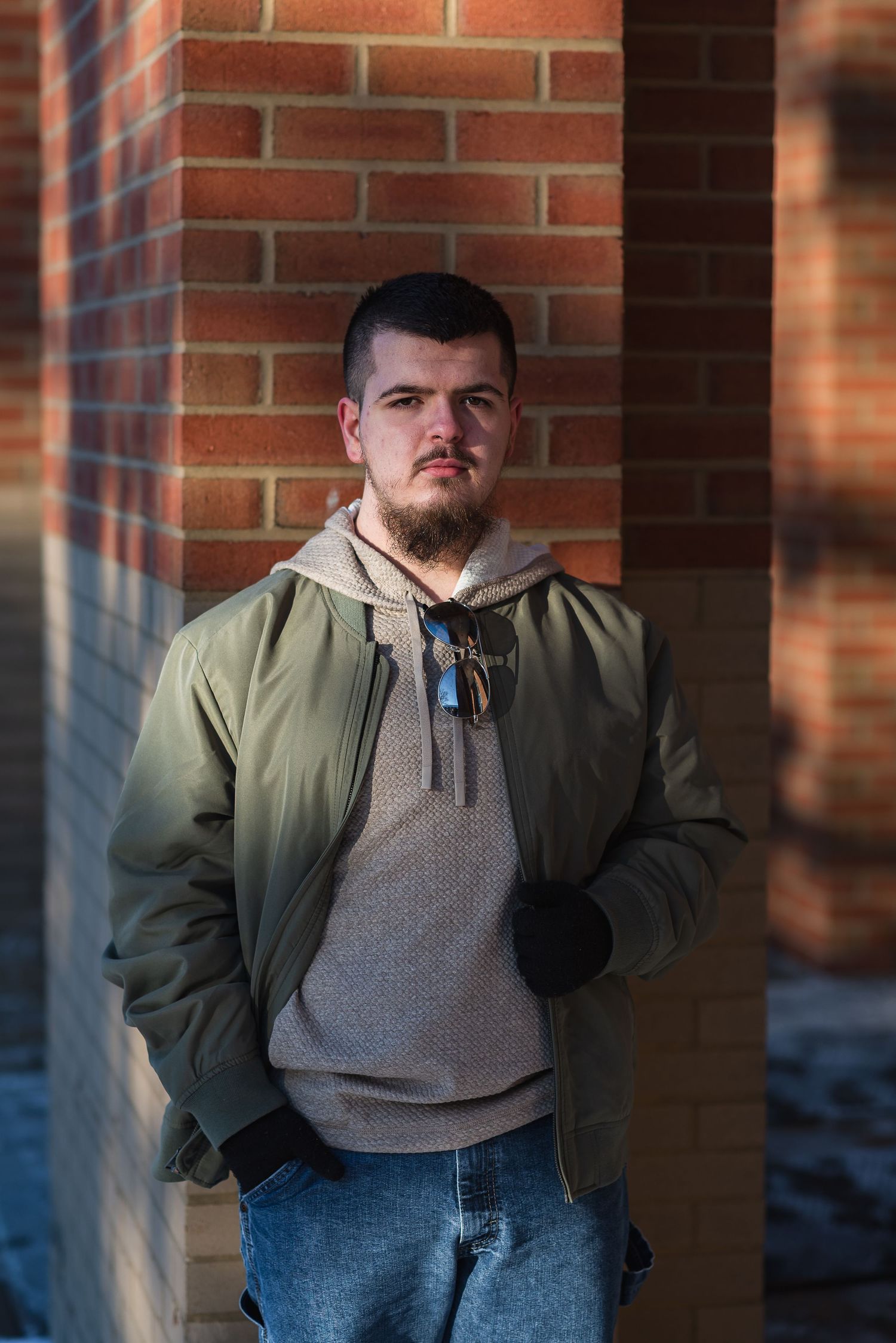Young man with beard in green bomber jacket and hoodie leaning against brick wall outdoors.