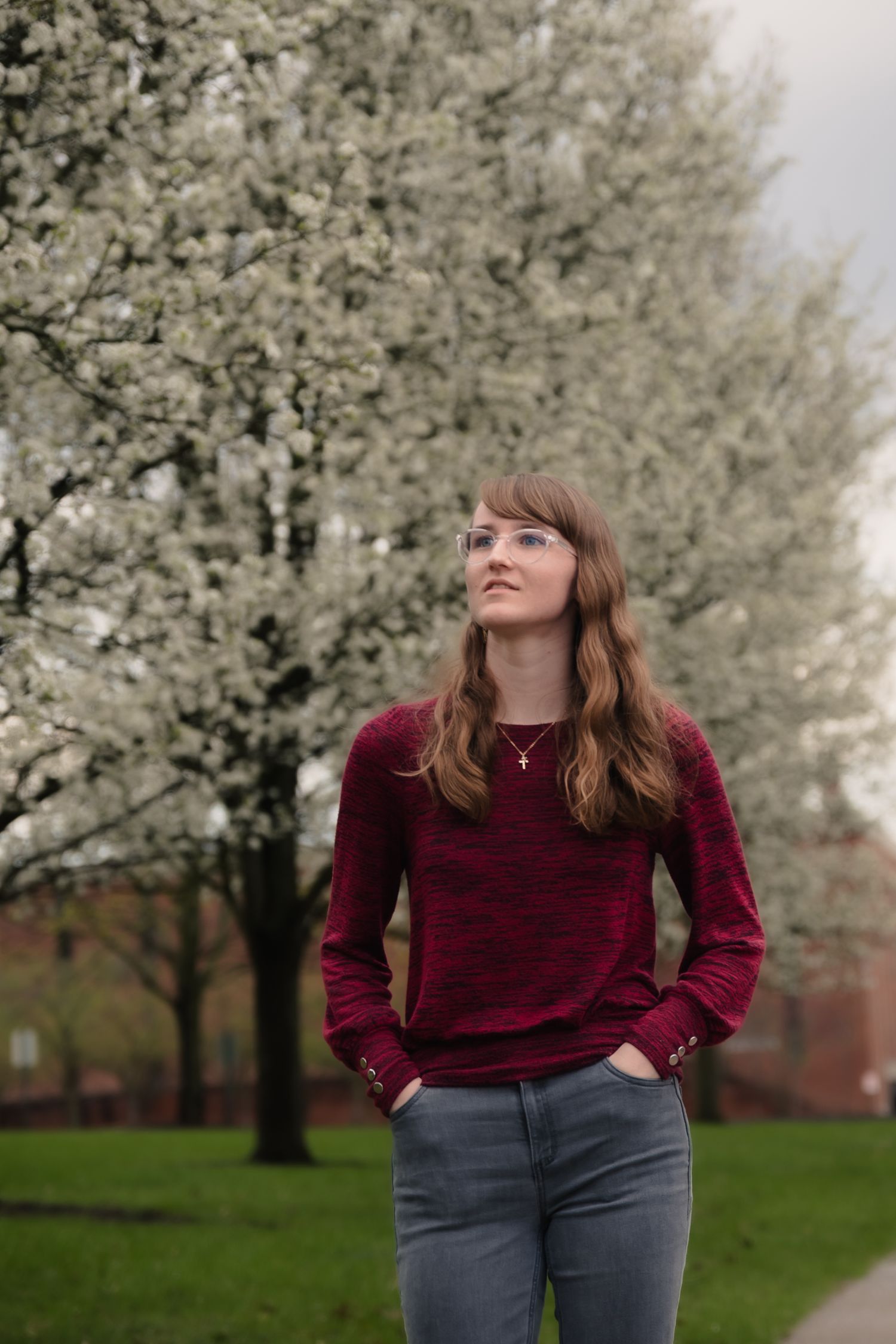 Young woman in red sweater and jeans stands before blooming white spring trees in a lush green park.