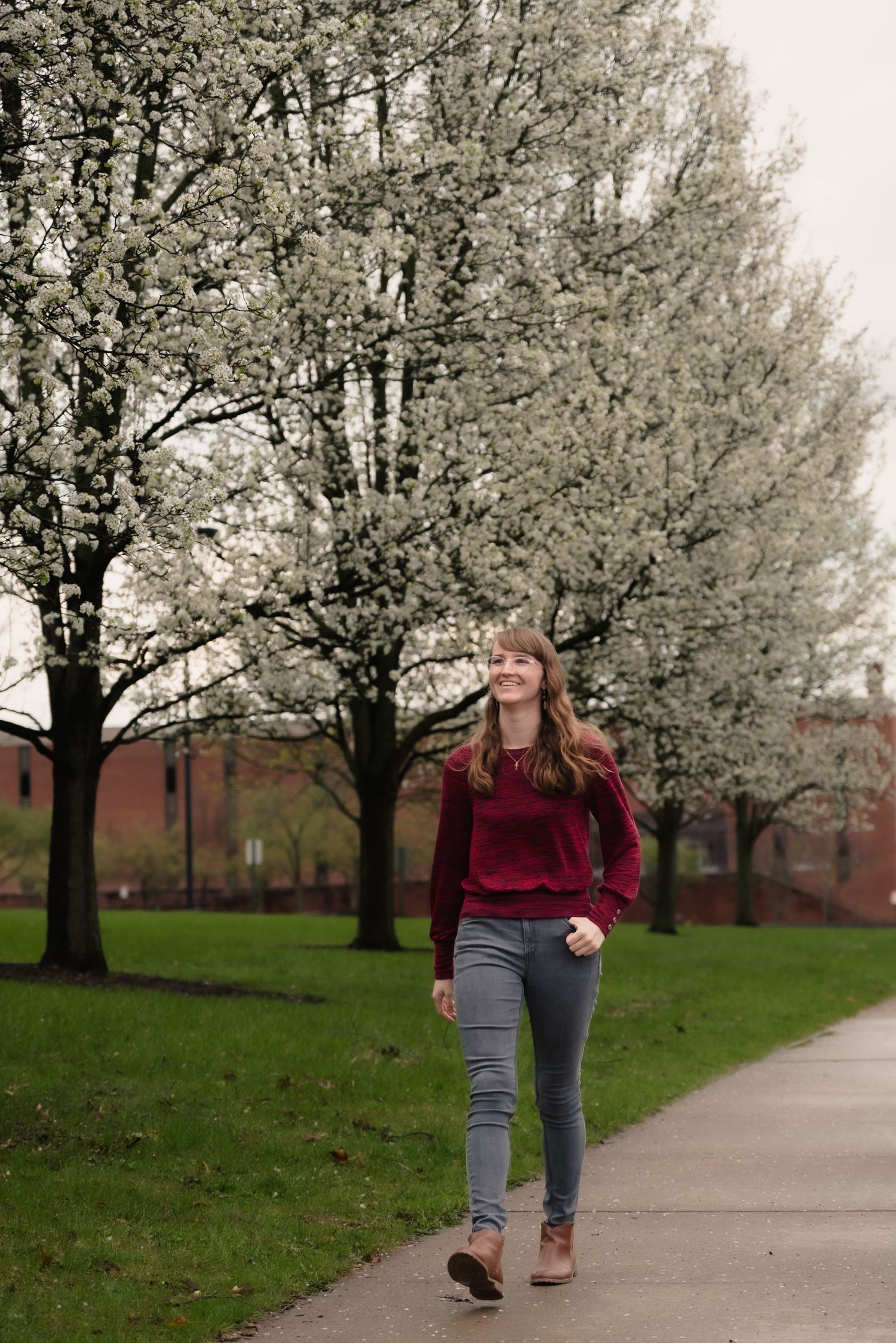 Young woman in burgundy sweater and gray jeans walking on a path beside blooming white spring trees in a park.
