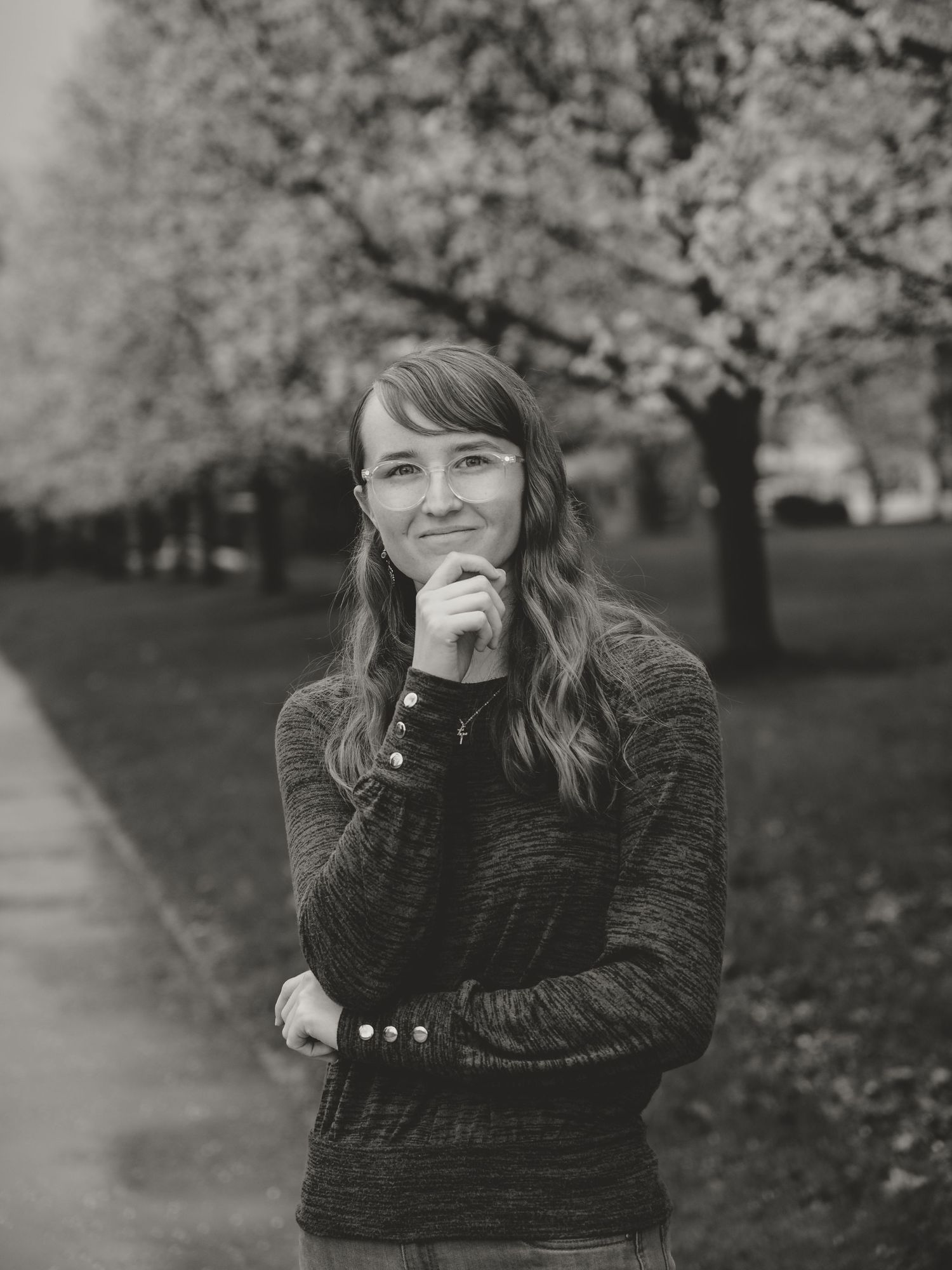 Young woman with glasses posing thoughtfully in a park with blooming trees in black and white photo.