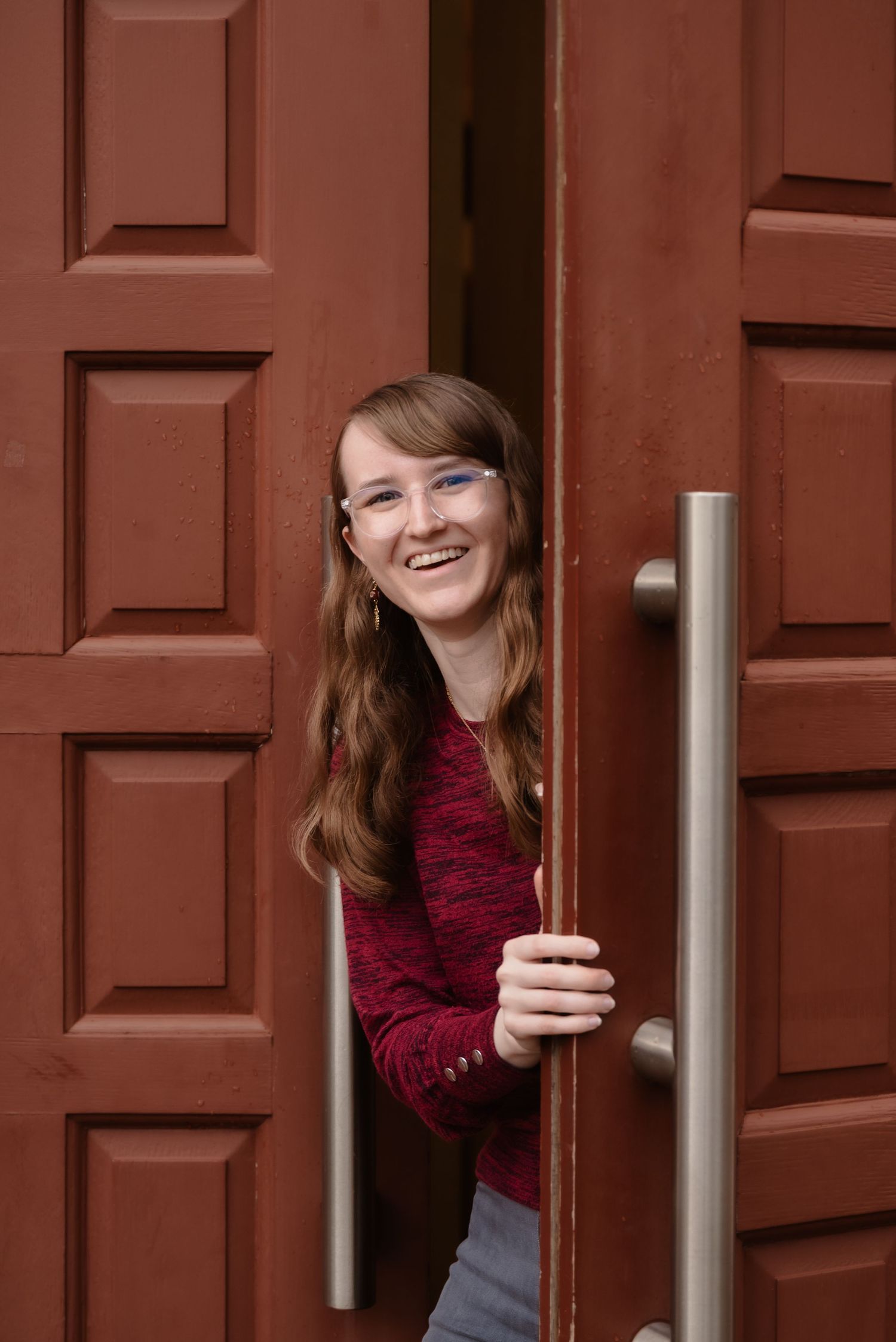 Smiling young woman with glasses peering through large red wooden doors, wearing a red top.