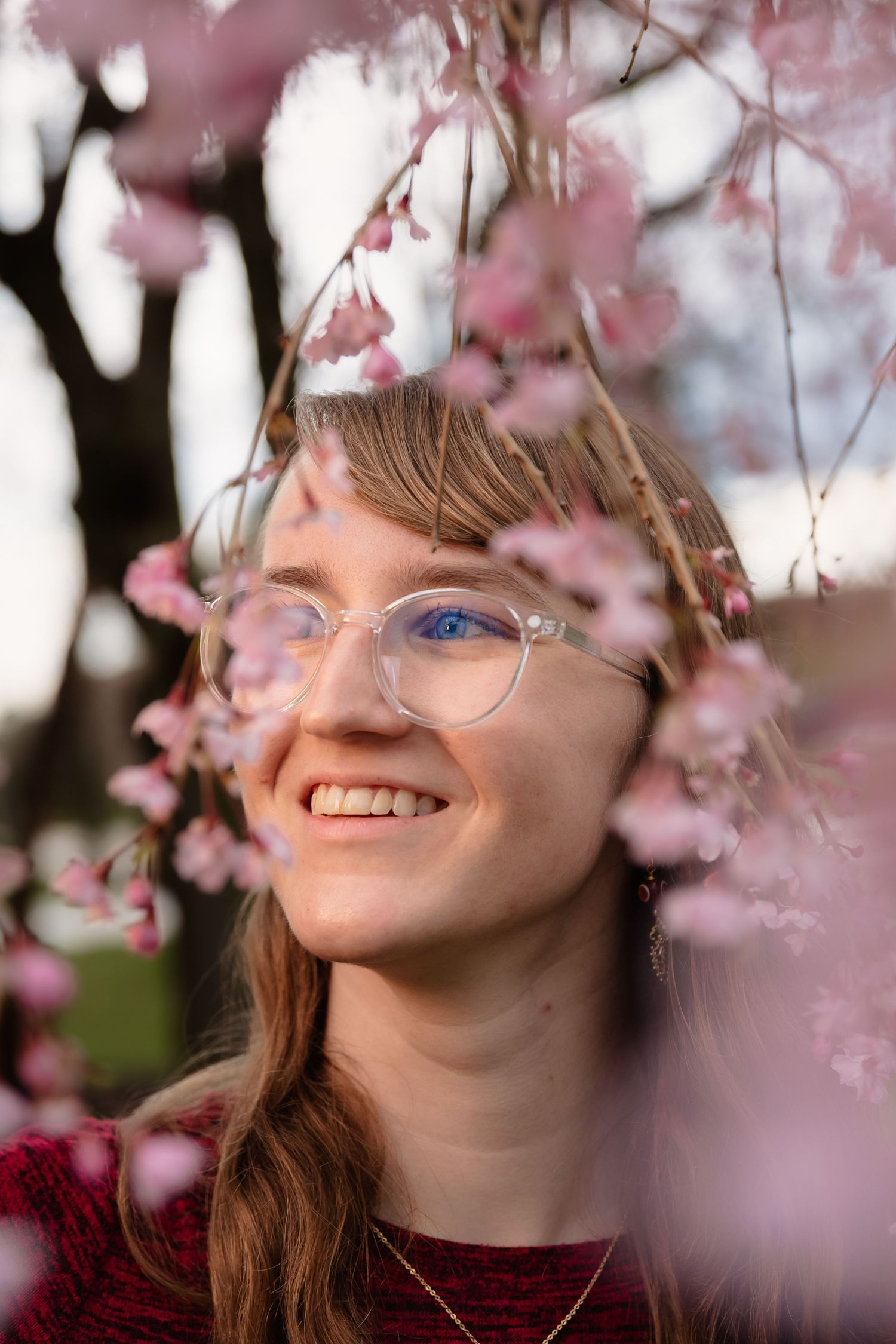 Smiling young woman with clear glasses surrounded by pink cherry blossoms in spring.