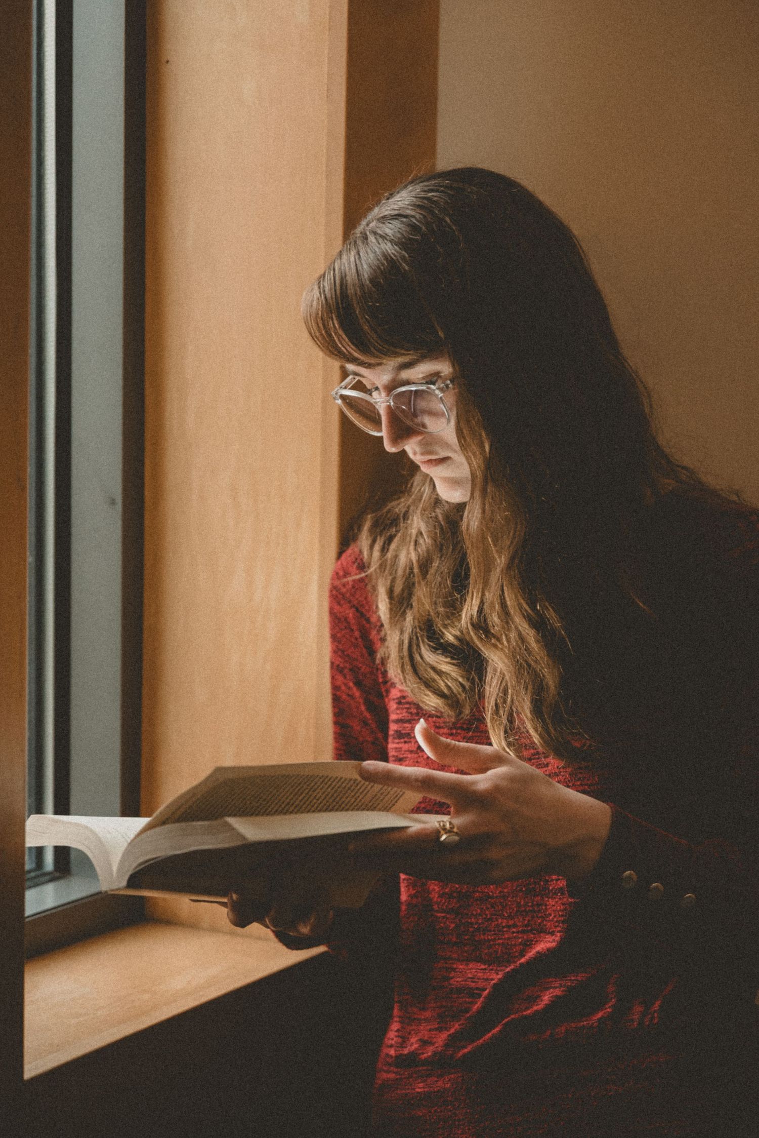 Young woman with glasses reading a book by a sunlit window, wearing a red sweater, with long brown hair.