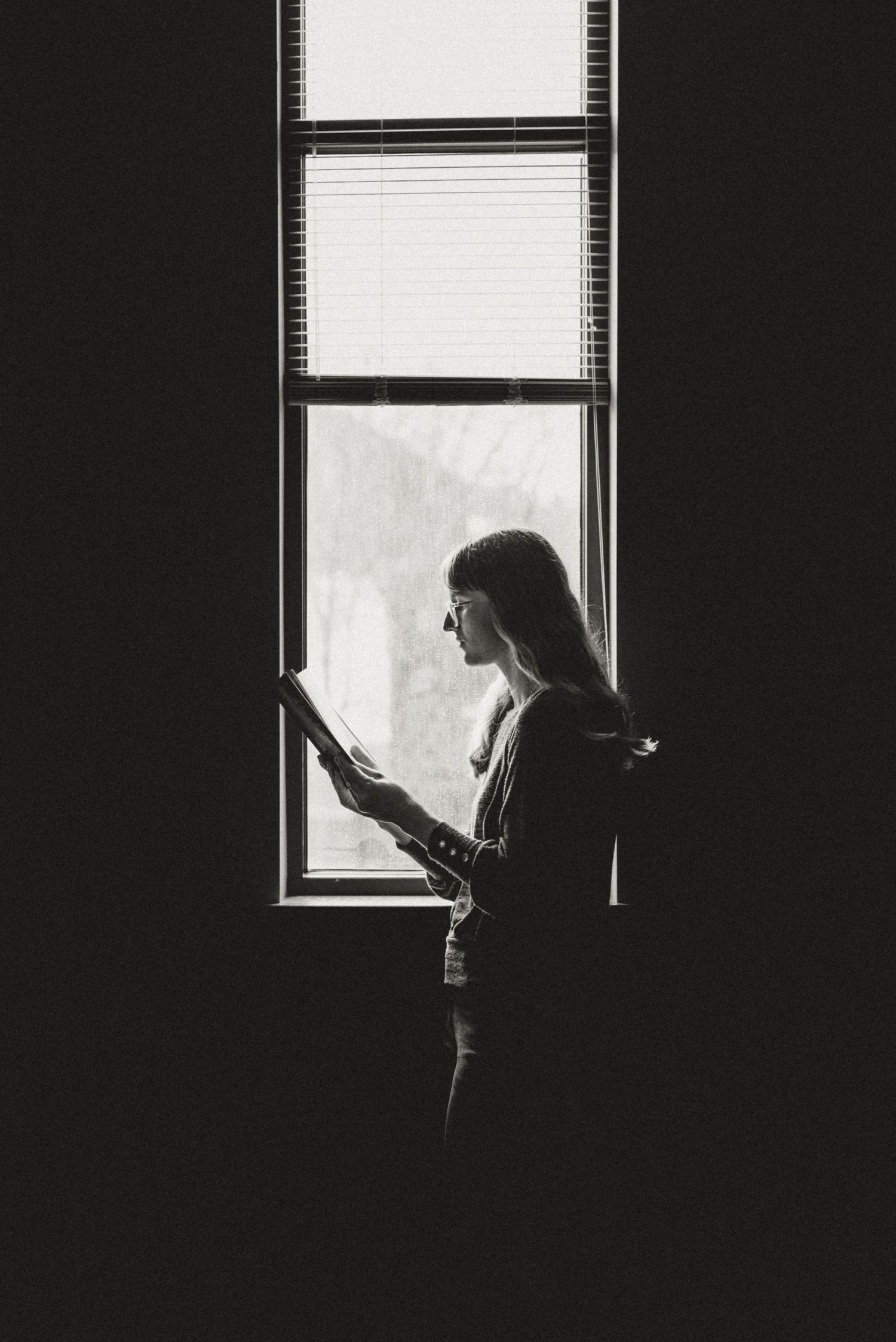 Black and white silhouette of a woman reading by a window with natural light streaming through blinds.