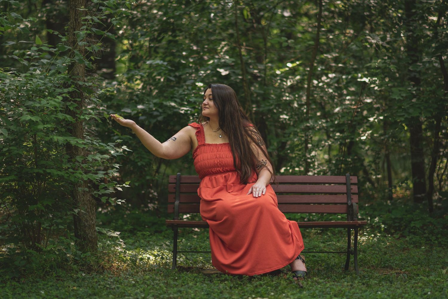 Graduate in red dress poses playfully in wooded outdoor setting.
