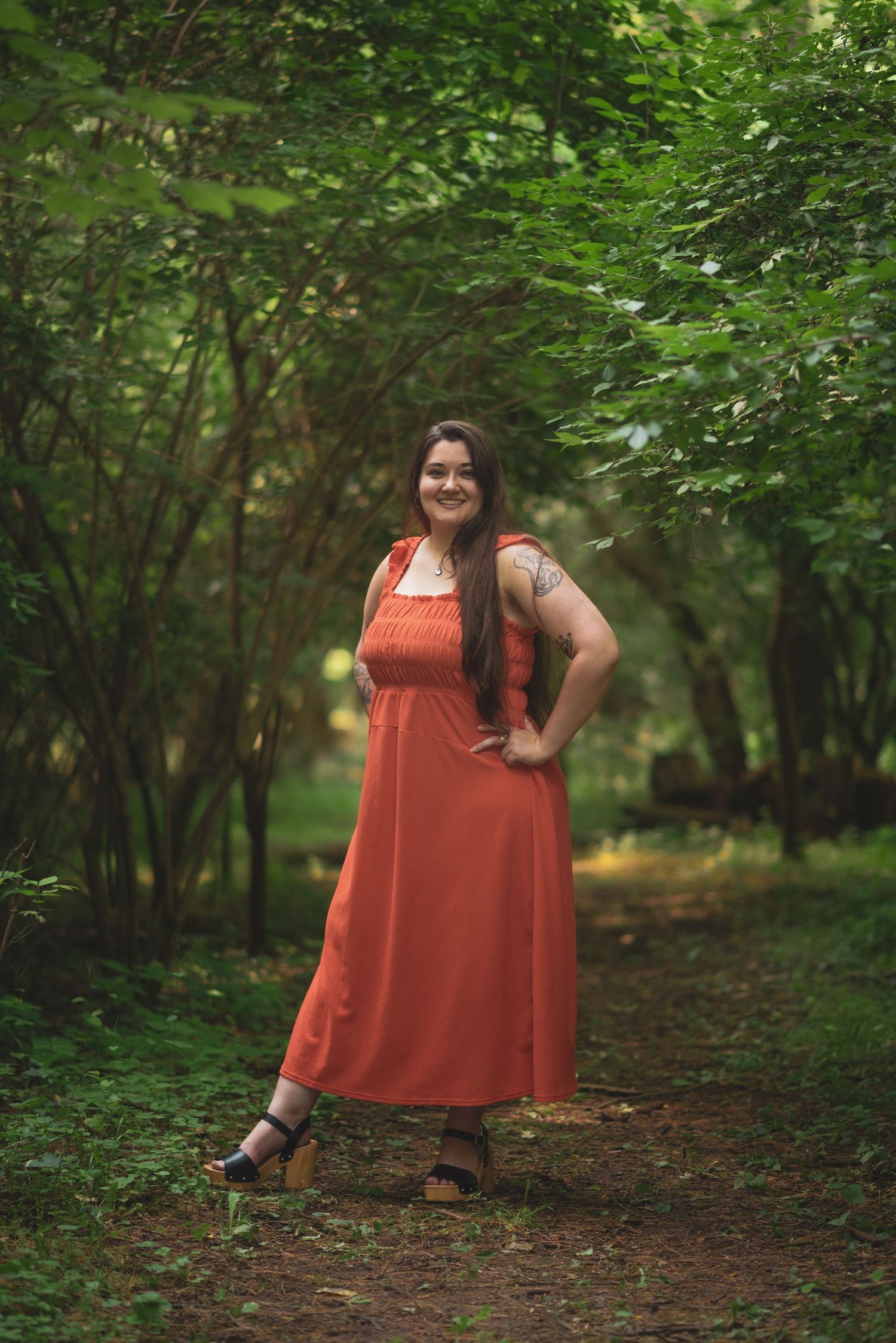 A person in a coral-colored dress poses confidently in a lush green forest setting.