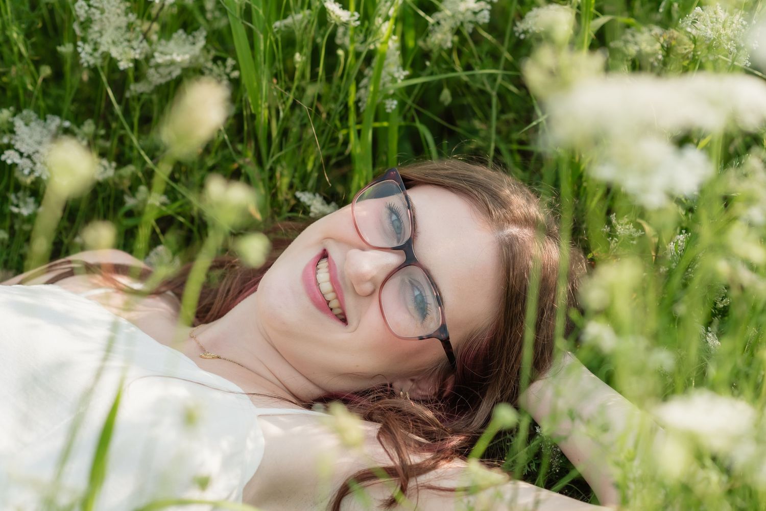 A smiling woman with glasses lying in a lush green meadow surrounded by white wildflowers on a sunny day.