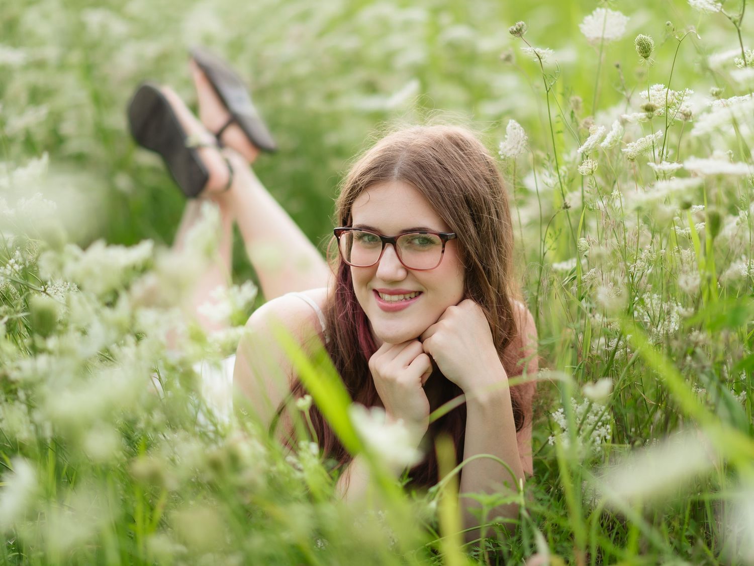 Young woman with glasses smiling while lying in a lush green meadow filled with white wildflowers.