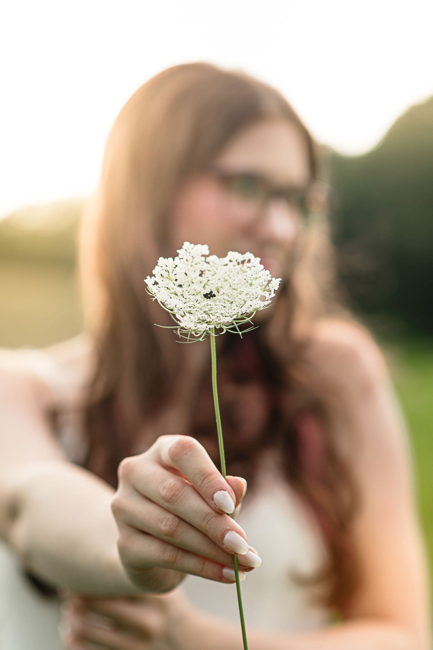 Young woman with glasses holding a delicate white Queen Anne's Lace flower toward the camera in golden sunlight.