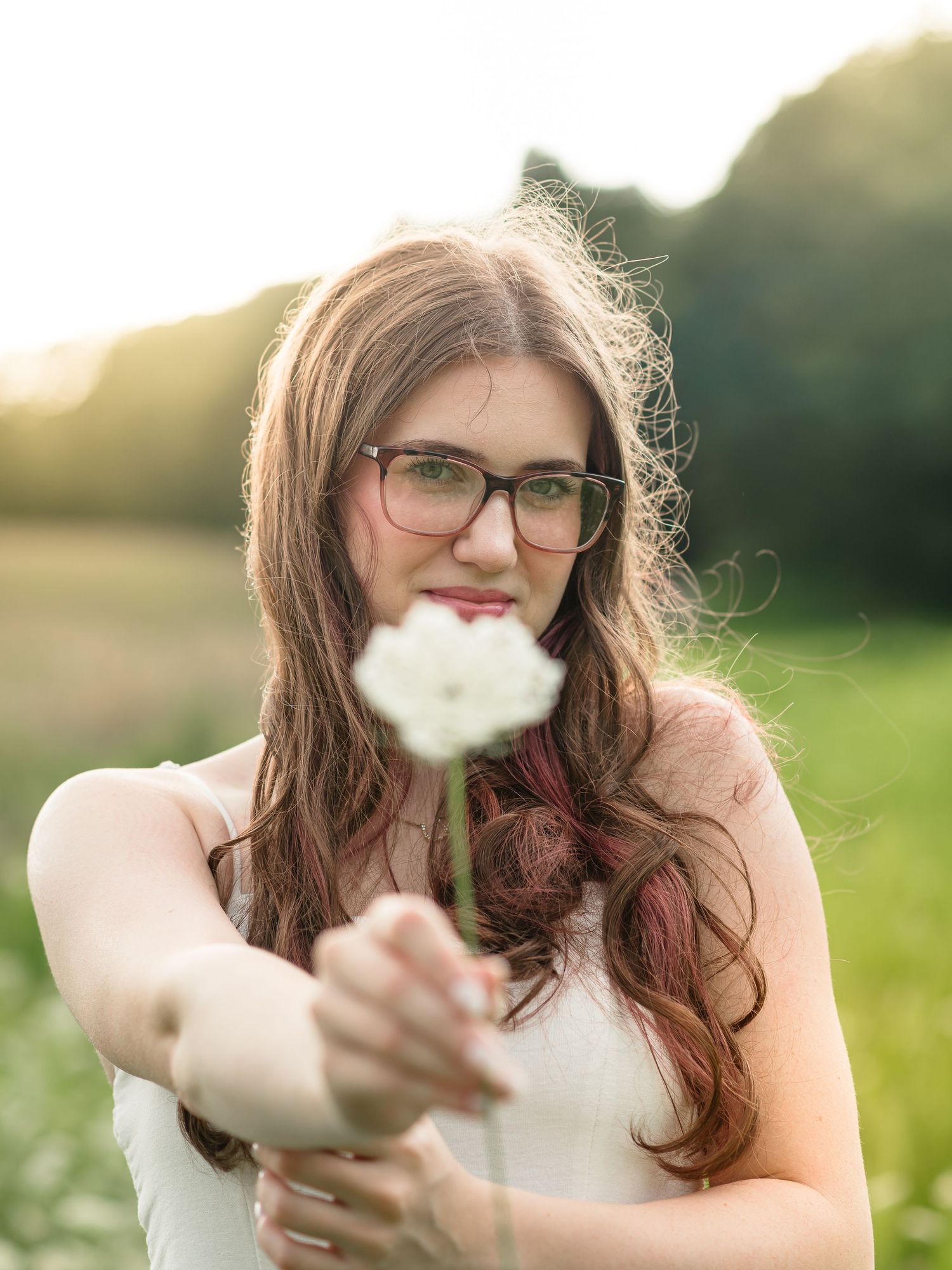 Young woman with glasses holding a white flower toward camera in a sunlit green field.