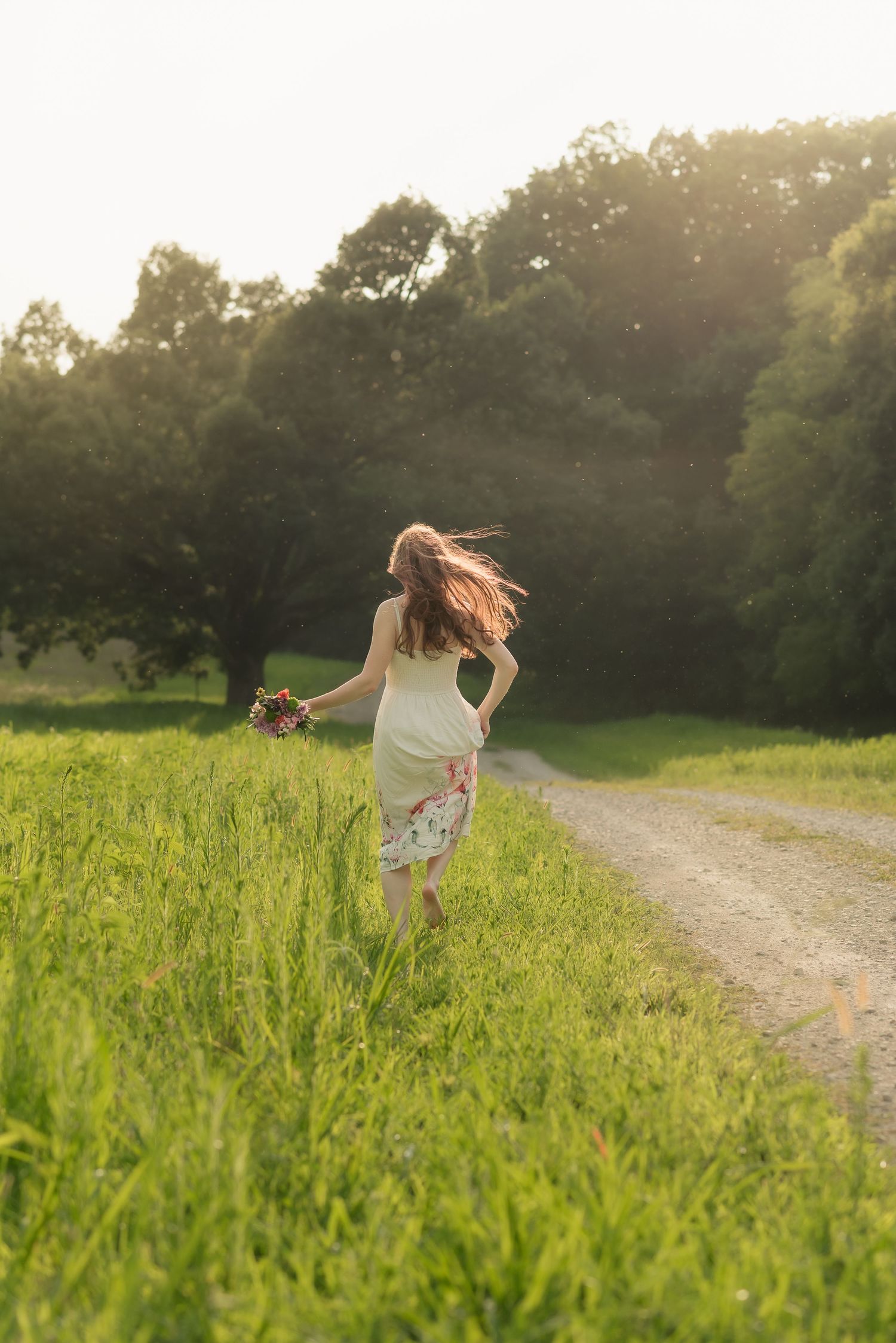 Woman in floral skirt running through green meadow holding wildflowers bouquet at golden sunset near dirt path.