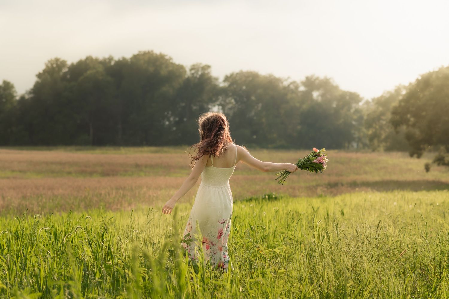 Woman in white dress walking through green field holding wildflowers with sunlit trees in background.