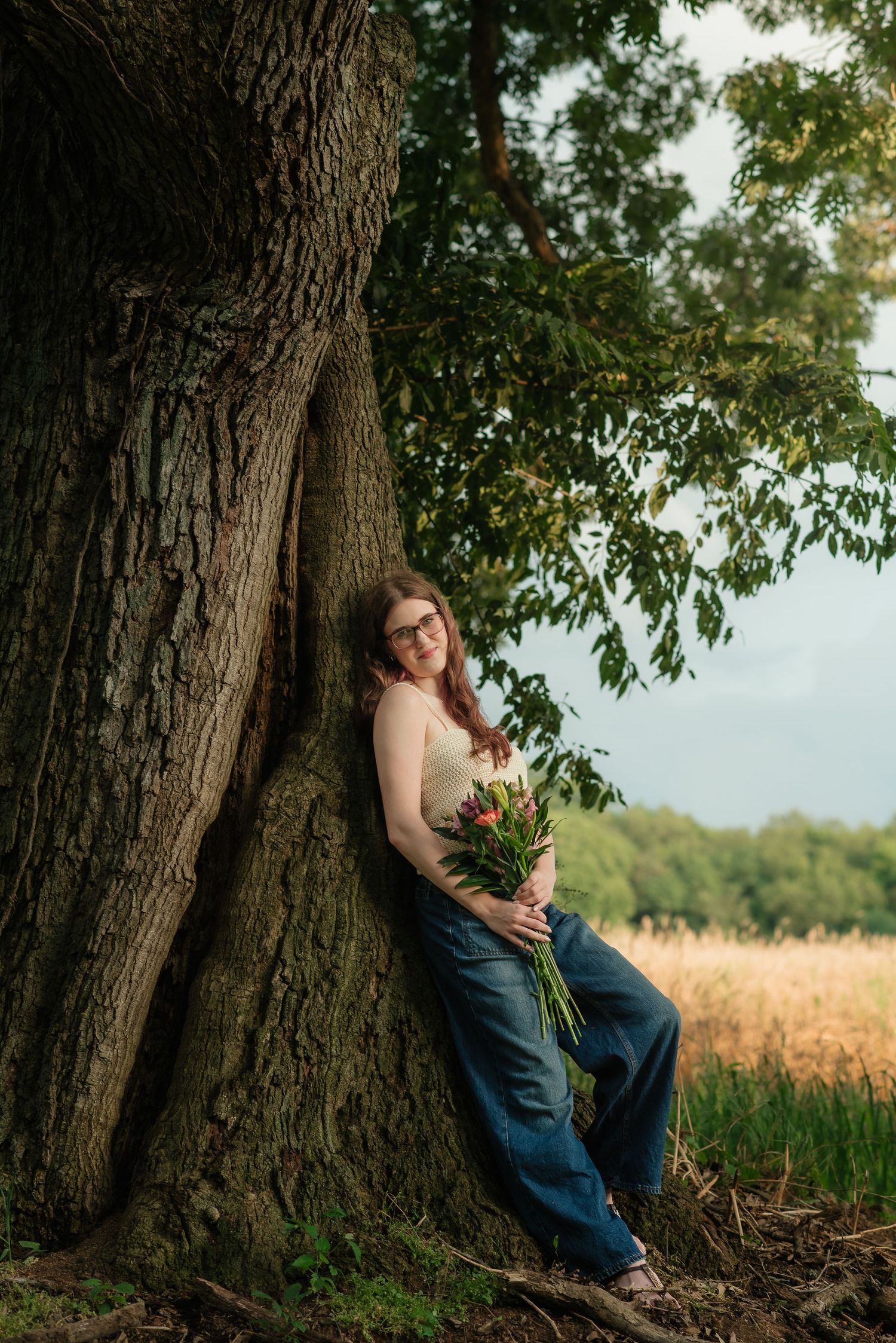 Young woman with glasses leans against large tree trunk holding colorful bouquet in jeans and tank top outdoors.