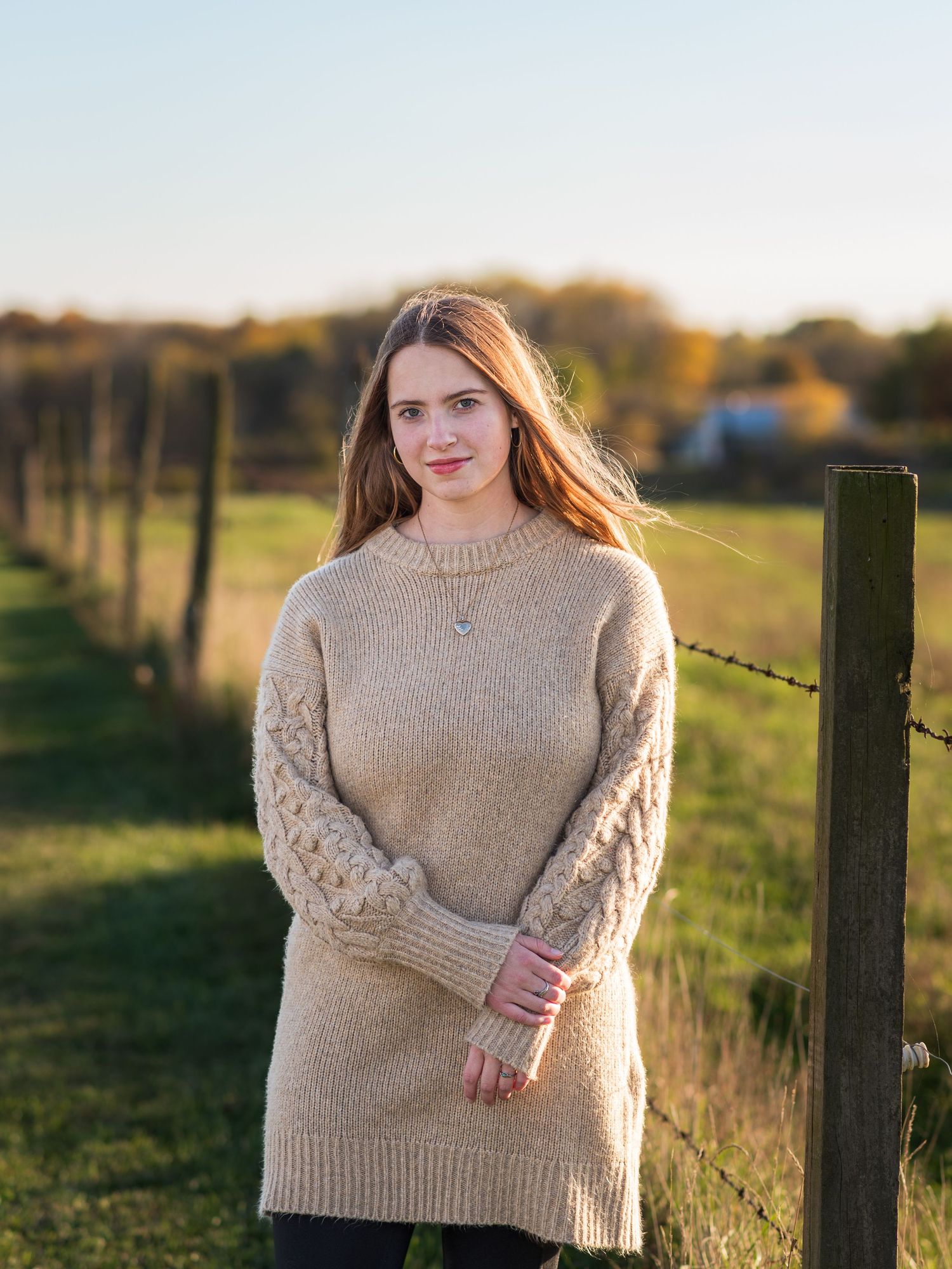 Woman wearing beige knit sweater poses in golden sunlight by rustic fence in countryside setting.