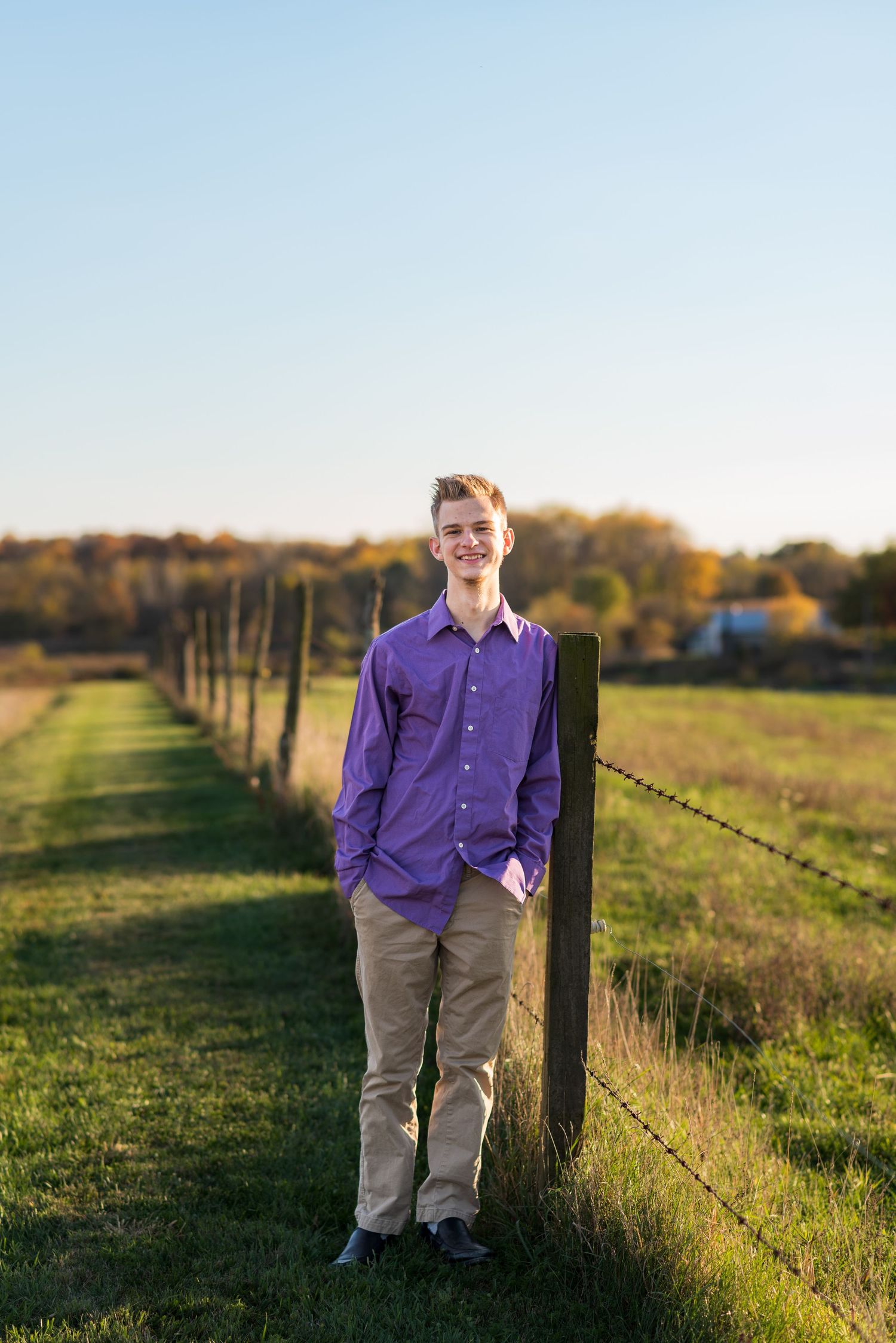 Someone in a purple shirt leans against a wooden fence post in a rural setting at sunset.