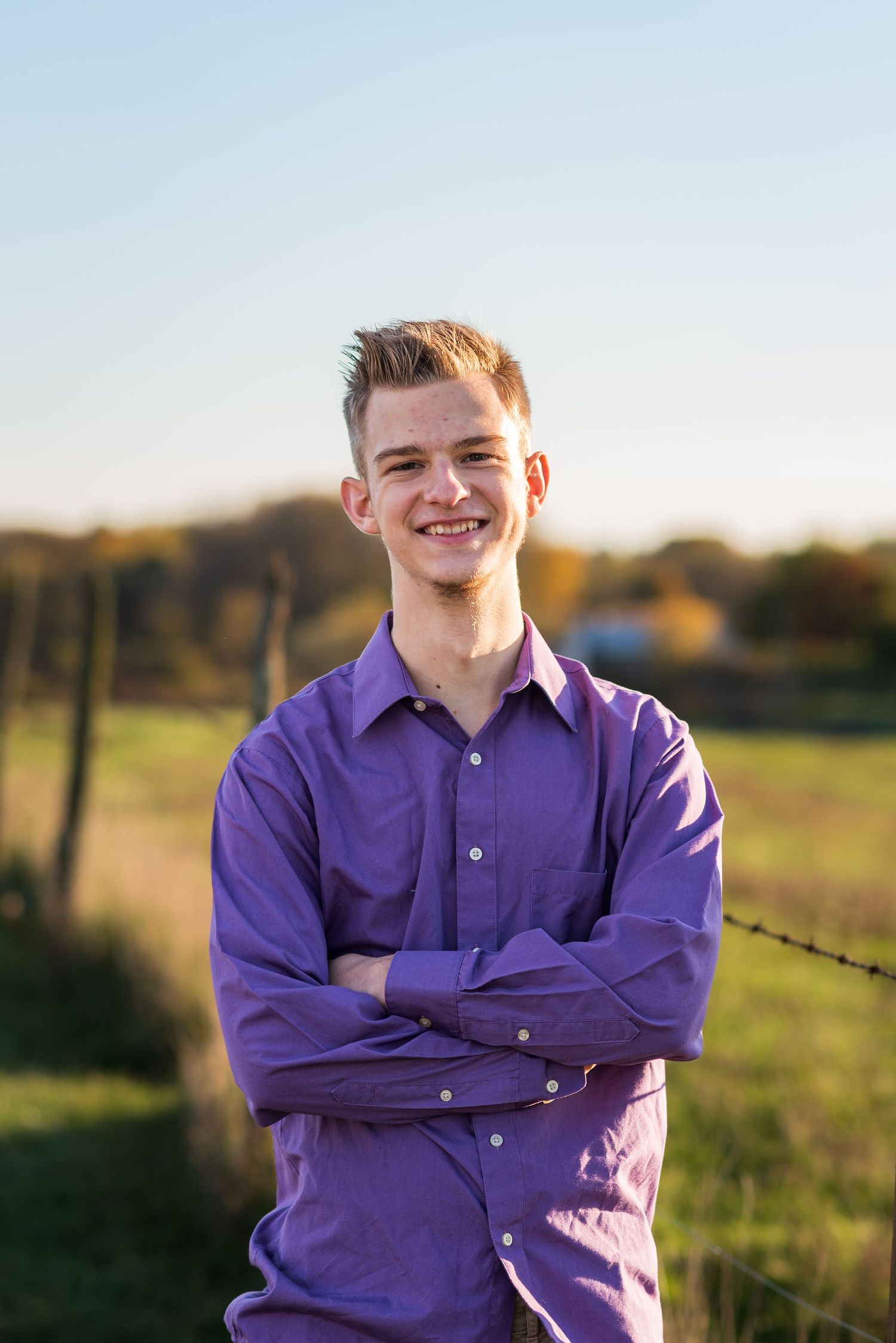 Person in purple button-down shirt poses confidently against rural fence at sunset.